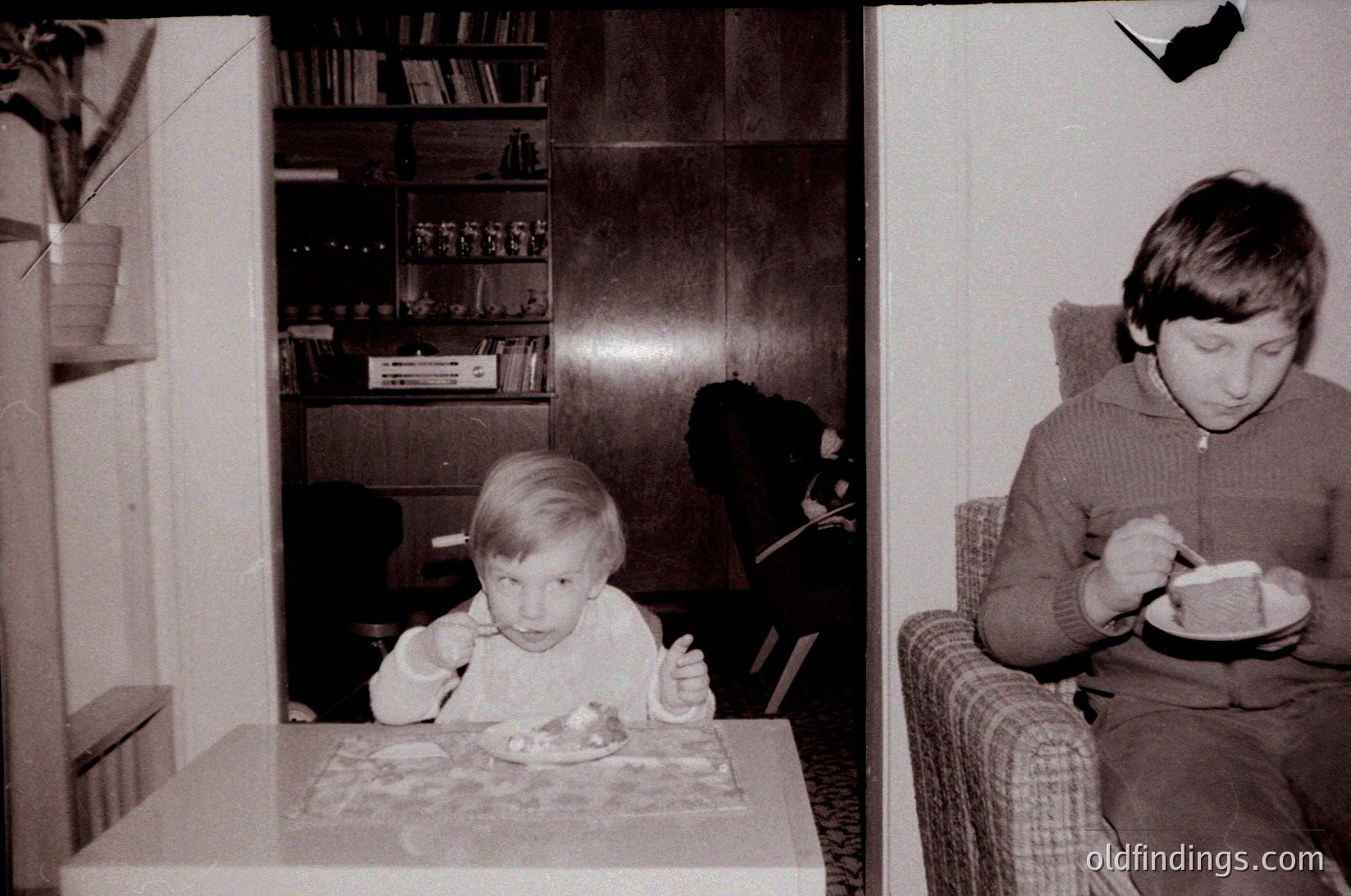 Two children eating indoors, likely mid-1970s. Child on left sits at a patterned tablecloth-covered table with a spoon, surrounded by bookshelves and a framed poster. Child on right in a plaid sweater holds a bowl, seated in a tufted armchair. Warm, domestic setting captures everyday family life.