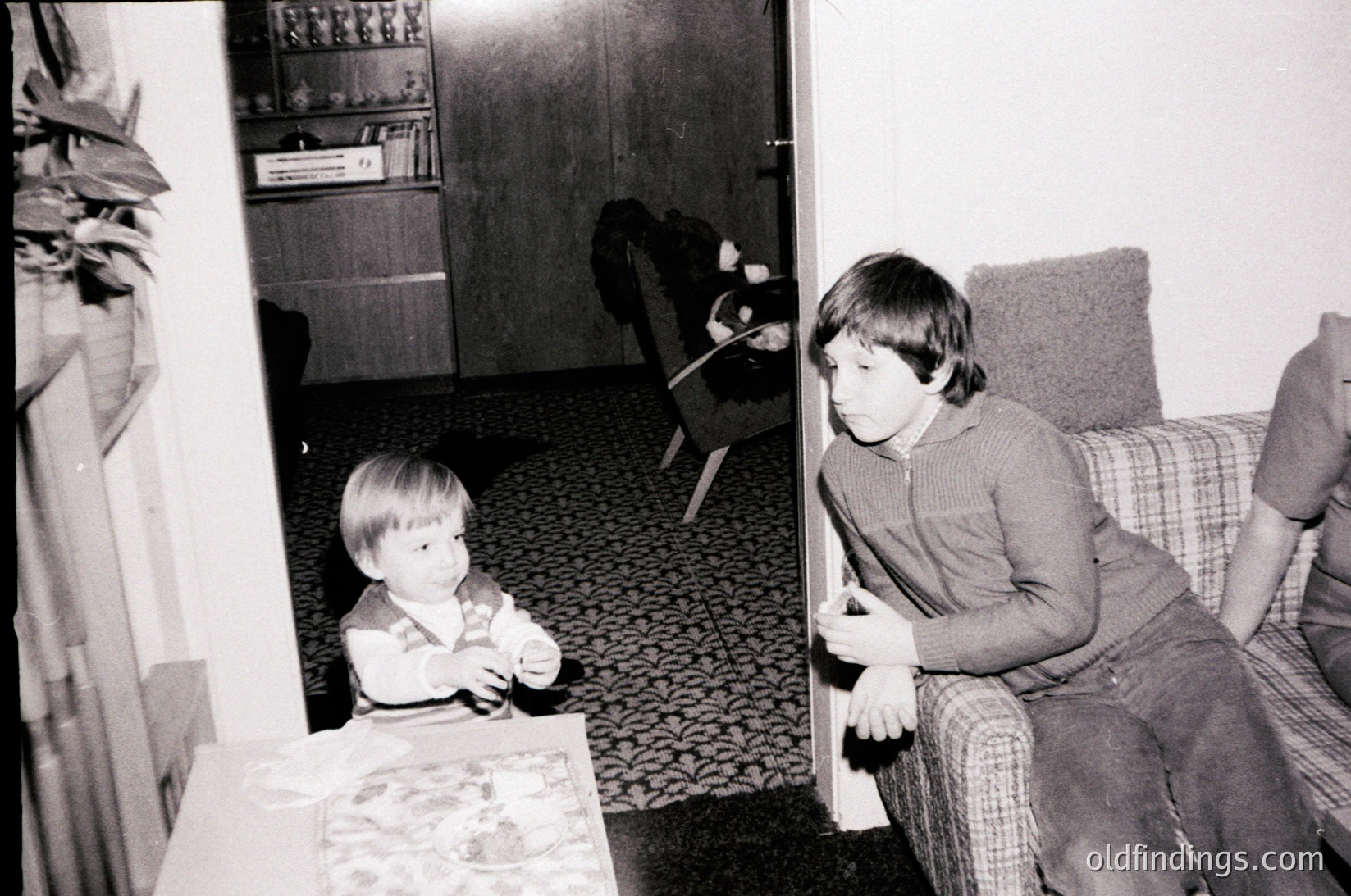 Two children in a mid-century living room, one seated on patterned sofa, the other standing by a low table with a patterned cloth. Bookshelf and armchair in background. Likely 1960s–1970s Western household.