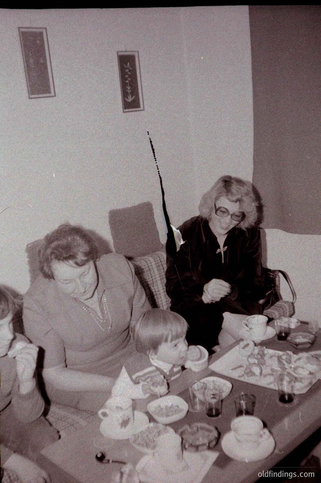 Vintage black-and-white indoor gathering featuring three adults and a child seated at a round table laden with teacups, cakes, and plates. Decor includes framed artwork and a patterned wall. Mid-century home setting, likely 1960s–1970s.