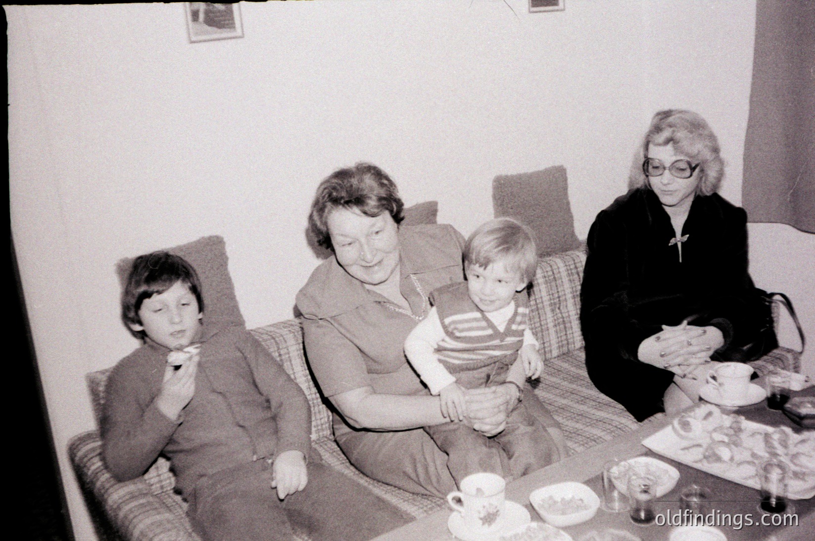 Black-and-white snapshot of a mid-20th century family gathering, likely 1960s–1970s. Three adults and a child seated on a patterned sofa, holding a baby. Table in foreground displays vintage teacups, saucers, and a plate of food. Warm, intimate domestic scene.