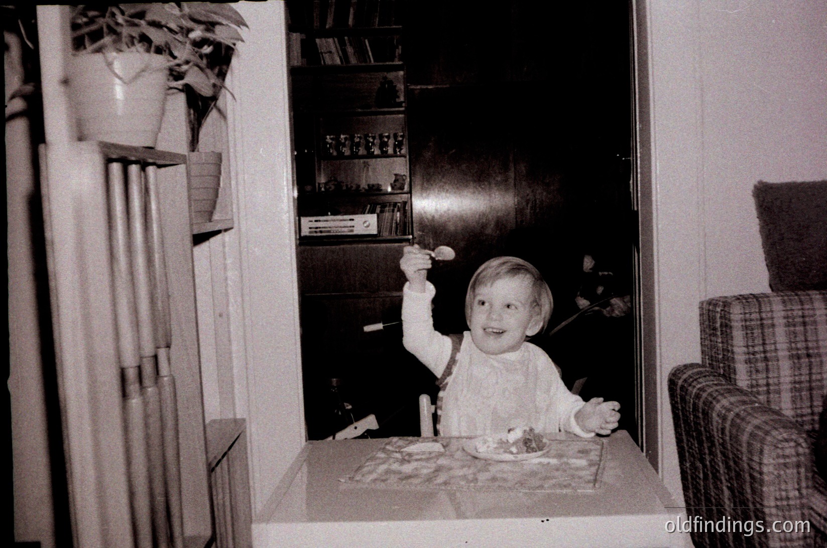 Vintage black-and-white photo of a child (approx. 2-3 years) seated at a small table, holding a spoon with cake. Mid-century home interior with wooden bookshelves, patterned tablecloth, and plaid armchair. Likely 1960s-1970s domestic setting.