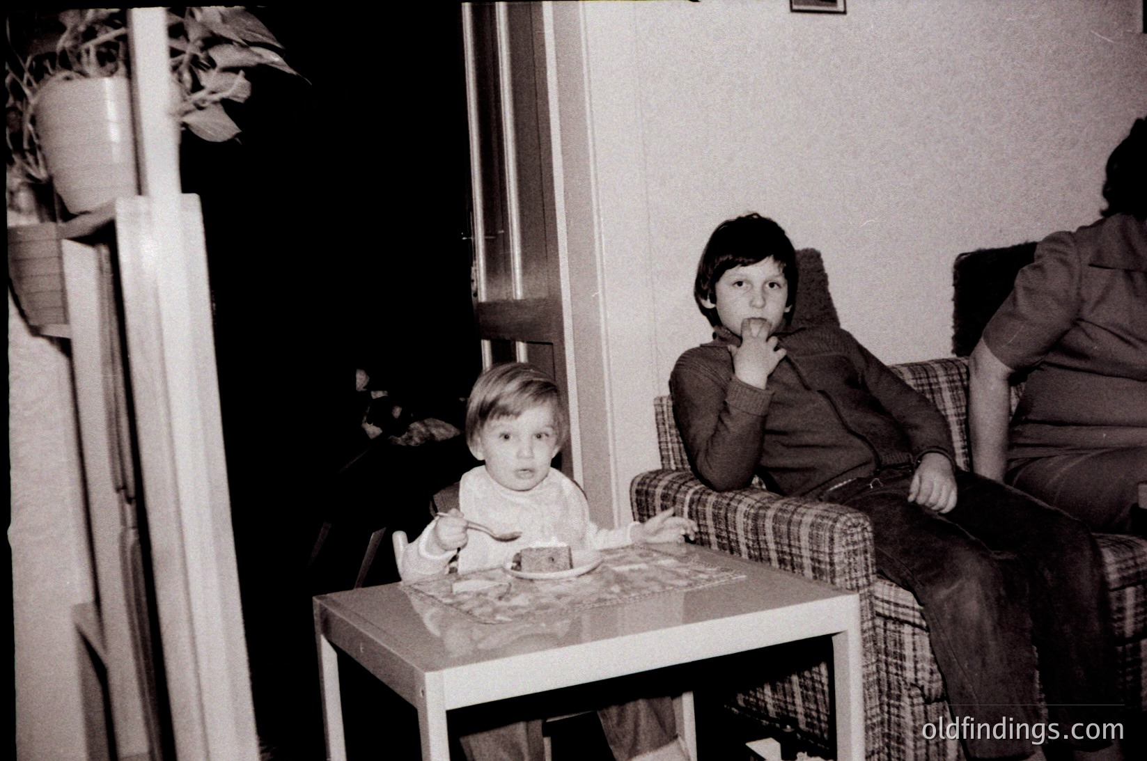 Two children seated indoors at a small table, likely mid-20th century. Infant in striped shirt holds a cake with lit candles; older child in plaid jacket covers mouth. Plain wooden furniture and minimal decor suggest a modest home setting. Candid, black-and-white snapshot captures a moment of celebration.