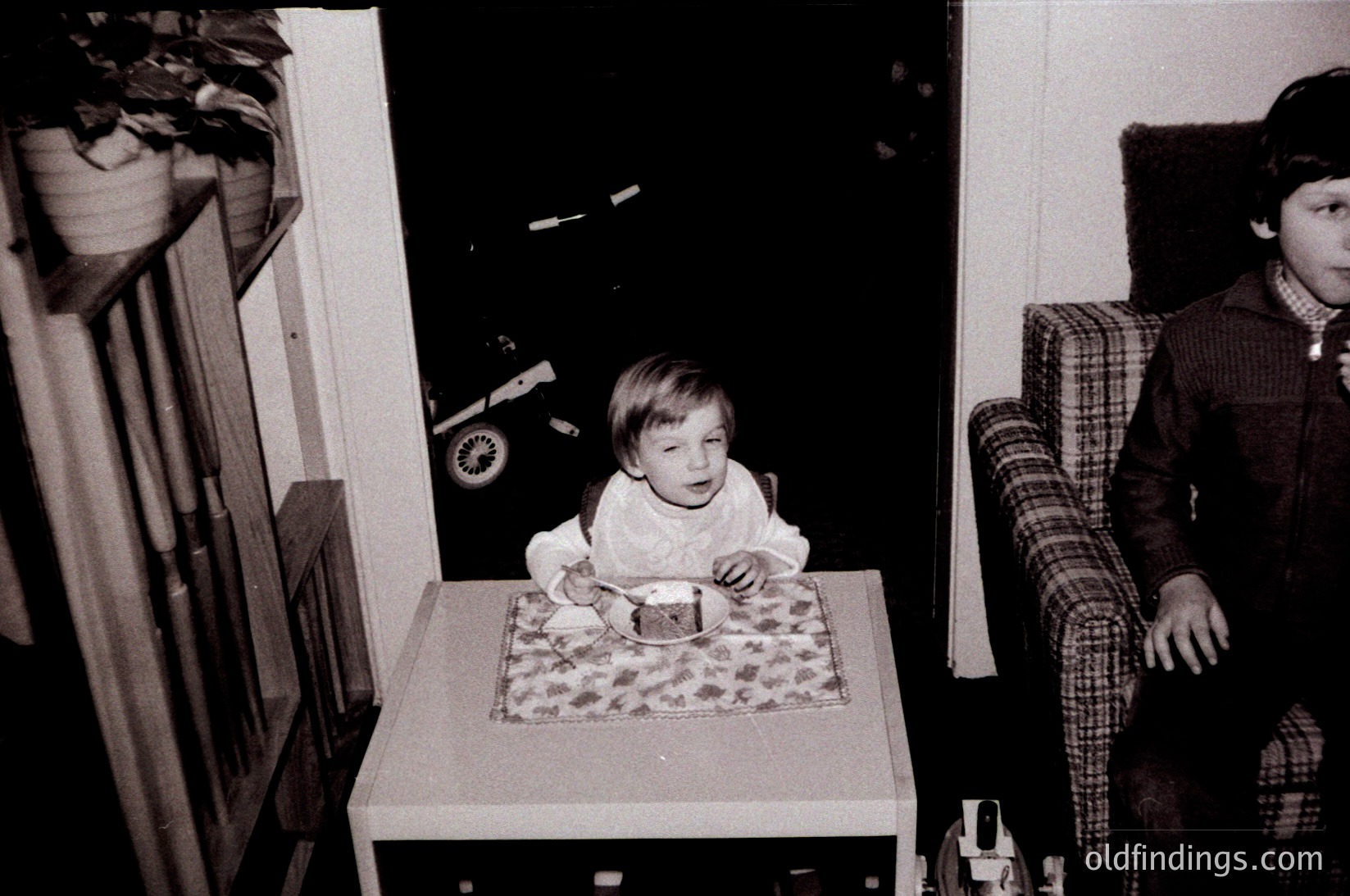 A toddler sits at a small wooden table in a mid-century interior, wearing a white long-sleeve shirt and patterned pants. Behind them, a child’s toy train rests on a shelf. A seated older child in a plaid shirt observes, while a vintage armchair and wooden cabinet frame the scene. Likely 1960s–1970s domestic setting.