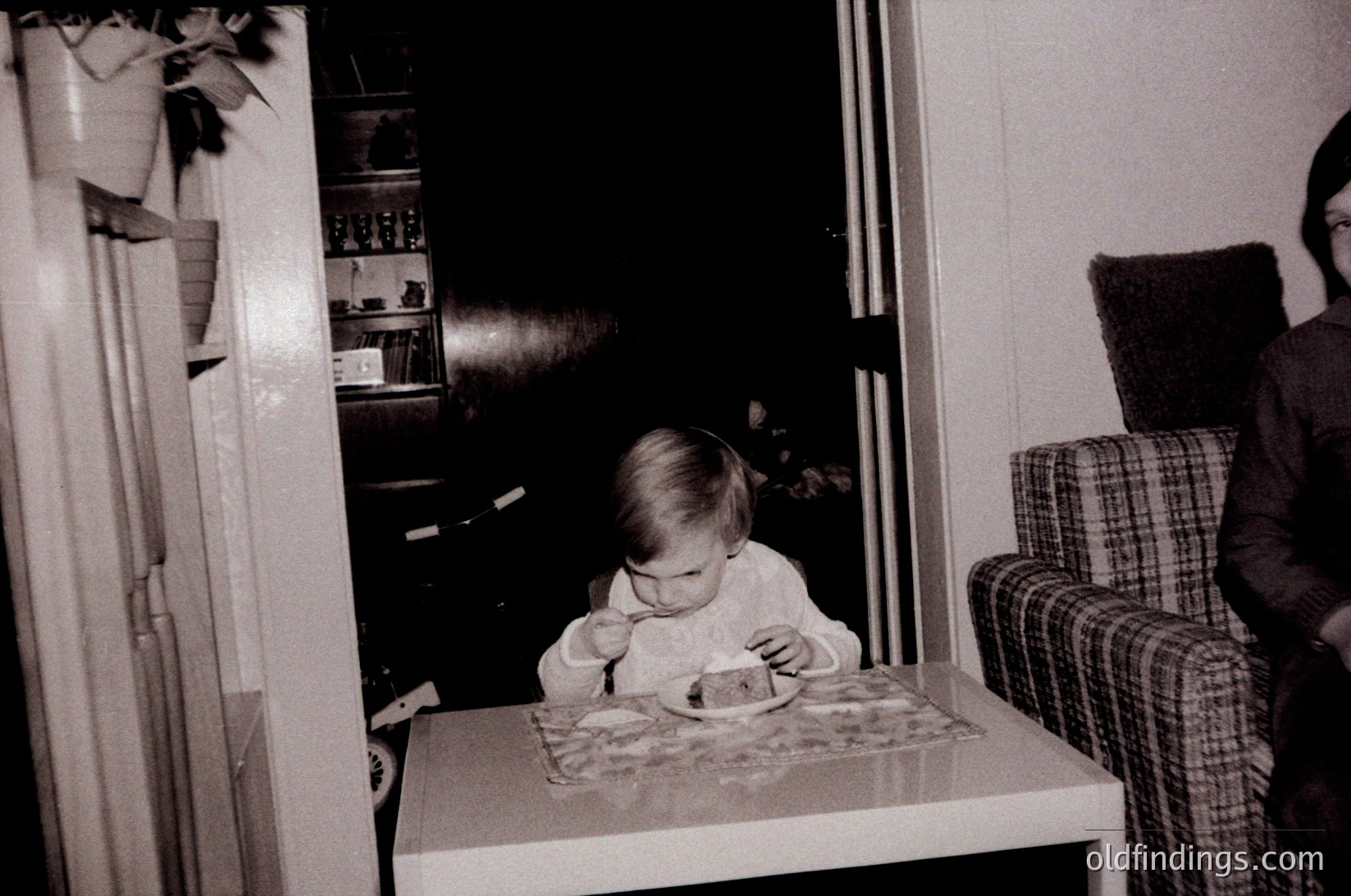 Vintage black-and-white photo of a toddler seated at a highchair, eating from a paper plate with a cartoon character. Mid-century kitchen interior with wooden cabinets, a checkered armchair, and a blurred adult figure in the background. Reflects 1960s-1970s domestic life.