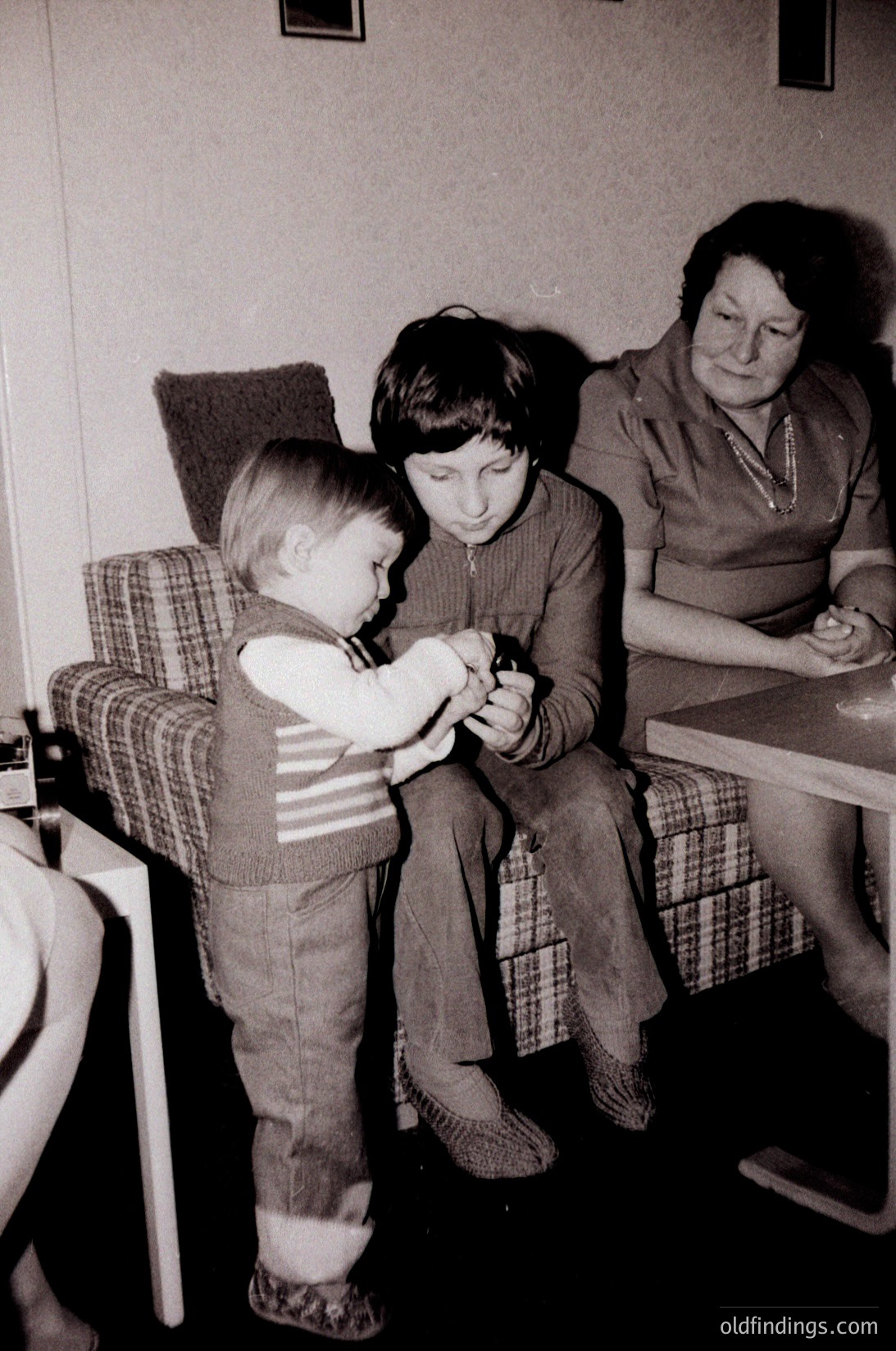 Vintage black-and-white indoor scene: a toddler in striped sweater and overalls kneels beside a seated older child in plaid pants, both examining a small object. An adult woman in a patterned blouse and apron sits nearby, holding a rolled-up paper. Mid-century furniture and simple decor suggest a 1960s–1970s domestic setting.