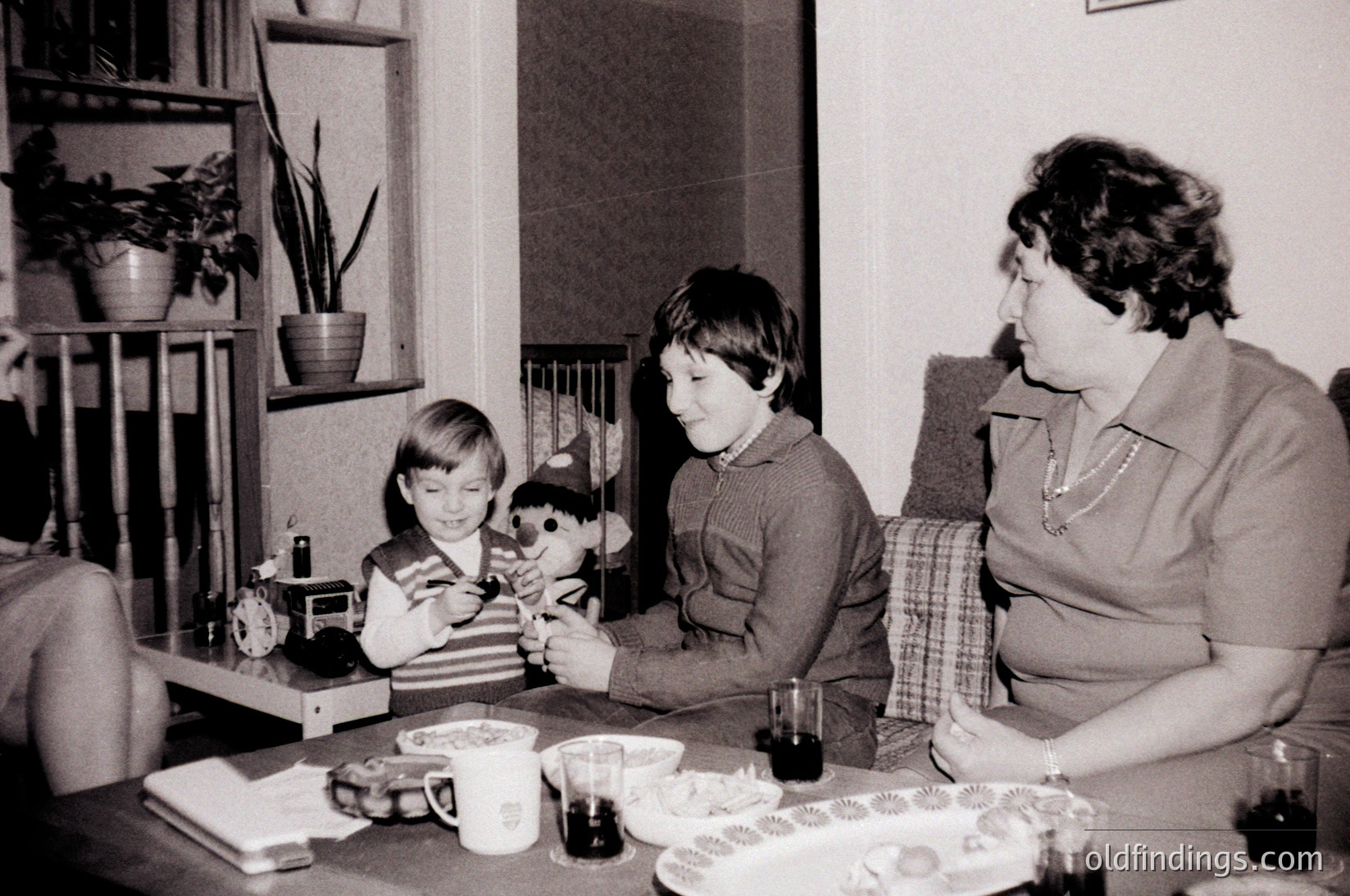 Three children and an adult share a mid-century indoor moment, likely 1960s–1970s. A boy in a sweater plays with a stuffed animal on a wooden table, while another child and woman observe. Vintage decor includes a potted plant, clock, and patterned plates. Warm, intimate family scene.