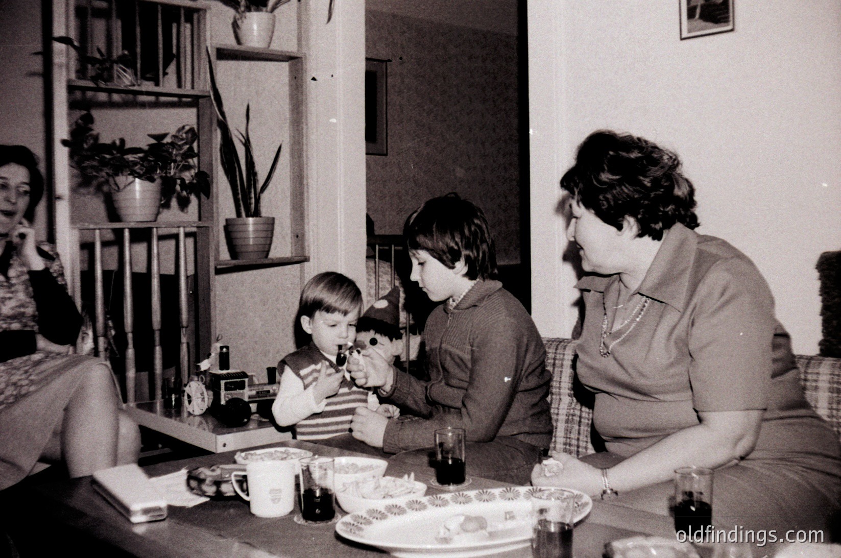 Vintage black-and-white indoor scene featuring a woman in a collared blouse and two boys at a table, likely mid-1970s. The woman holds a camera, capturing the younger boy’s reaction to a toy train set. Decor includes potted plants, a clock, and a coffee table with glasses and a plate of cookies. Warm, intimate family moment.
