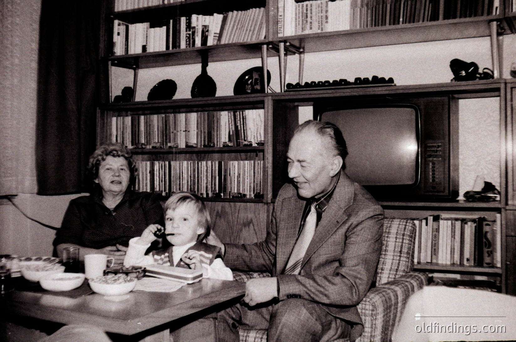 A mid-20th-century indoor family moment: elderly couple and child seated at a small wooden table, surrounded by bookshelves filled with volumes and a vintage TV. Classic 1960s-70s home decor with patterned upholstery.