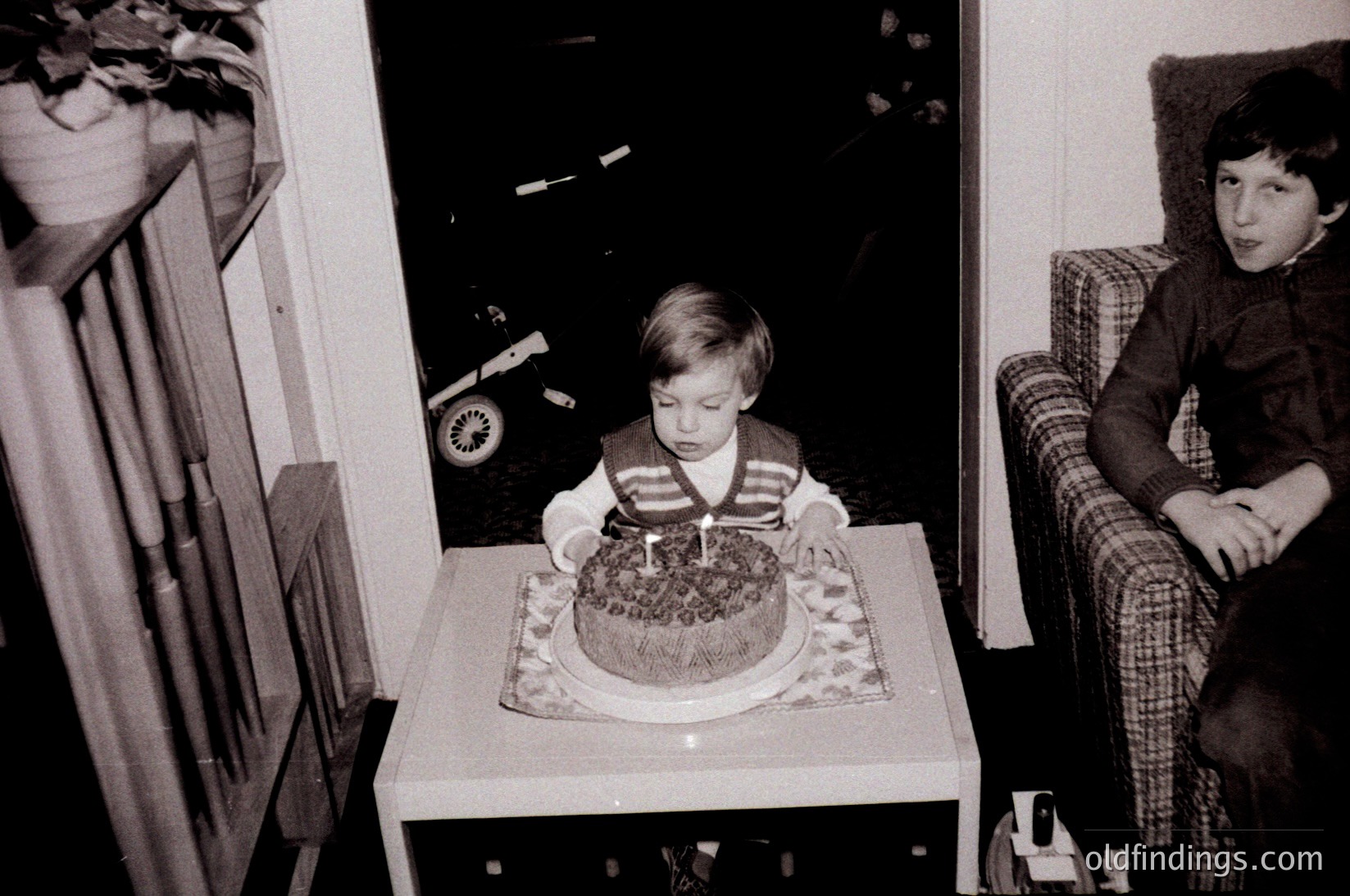 Vintage black-and-white indoor scene: young child seated at a small table blowing out a single candle on a decorated sheet cake, surrounded by mid-century home decor. Older child in plaid sweater sits on a chair beside. Toy car and patterned curtains add nostalgic 1960s–1970s charm.