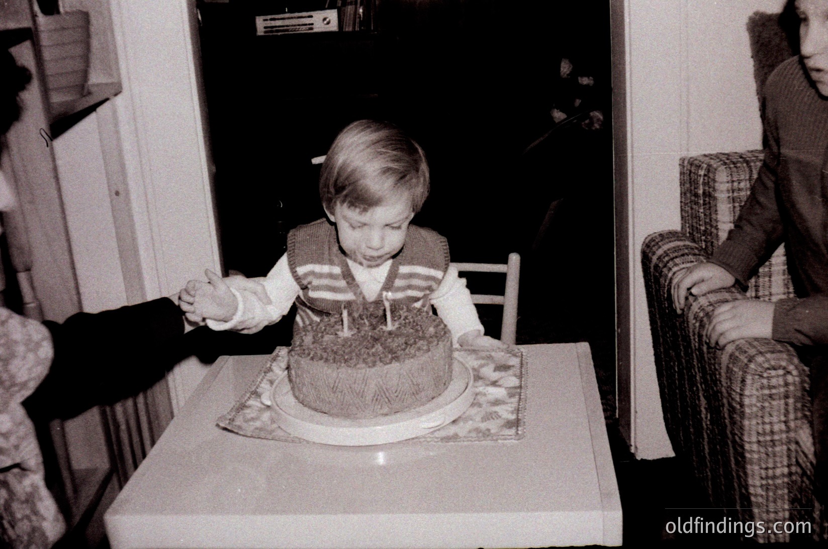 Young child blowing out candles on a round chocolate cake, seated at a small table in a mid-century indoor setting. Patterned tablecloth and plaid armchair suggest 1960s–1970s domestic decor. Warm, intimate family moment captured in black-and-white.