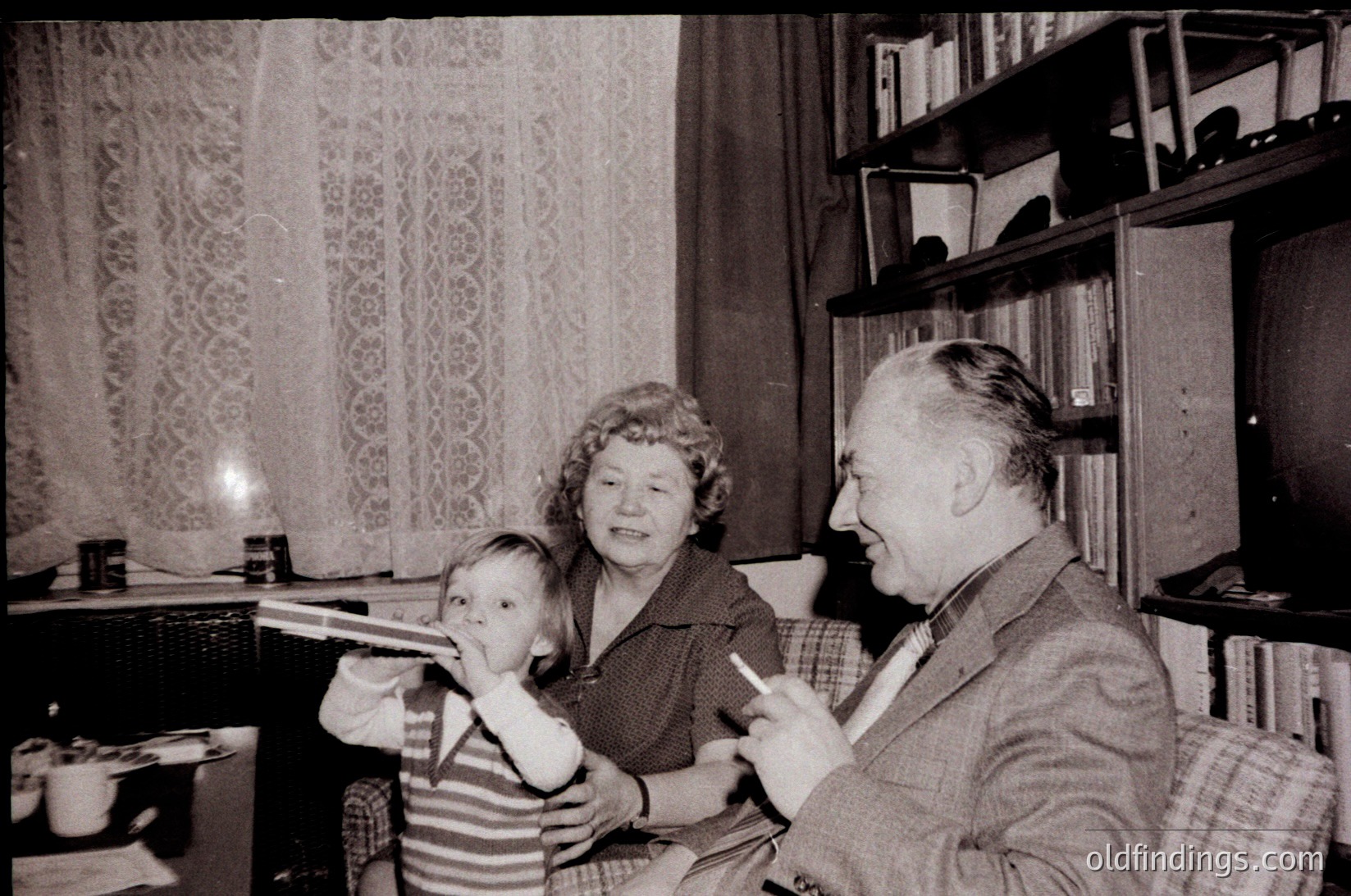 Mid-20th century (1950s–60s) indoor family moment: elderly couple and child in a cozy living room with patterned curtains, wooden shelves, and a vintage radio.