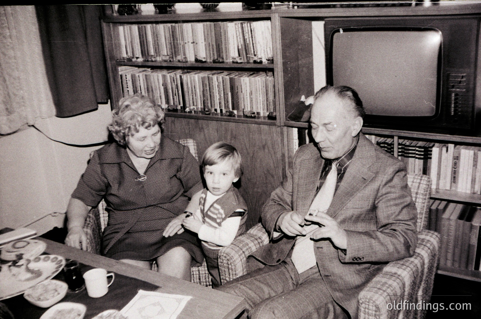 Mid-20th century family moment in a cozy living room: elderly couple and child seated on a sofa beside a coffee table with teacups. Bookshelves filled with bound volumes flank a vintage TV. Formal attire suggests a special occasion or weekly gathering.