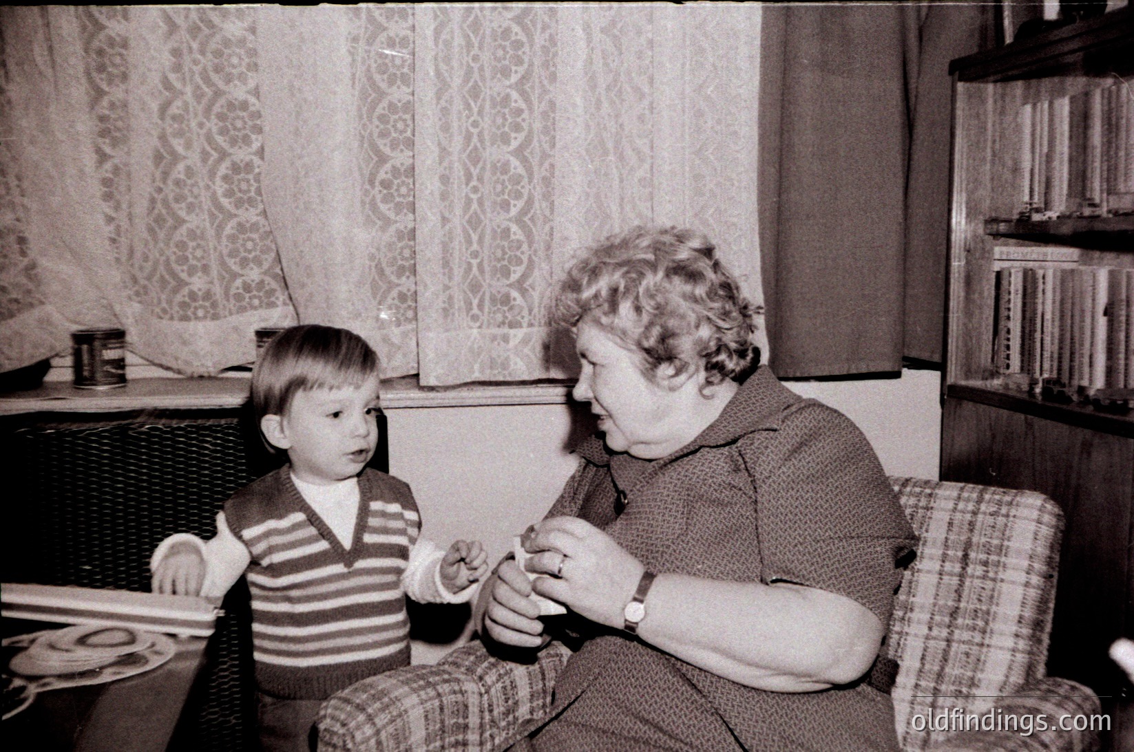 Vintage black-and-white photo of an adult woman and young boy engaged in a craft activity indoors, likely mid-20th century. Woman holds scissors, boy observes closely. Patterned curtains and bookshelf with vinyl records in background suggest a mid-century home setting.