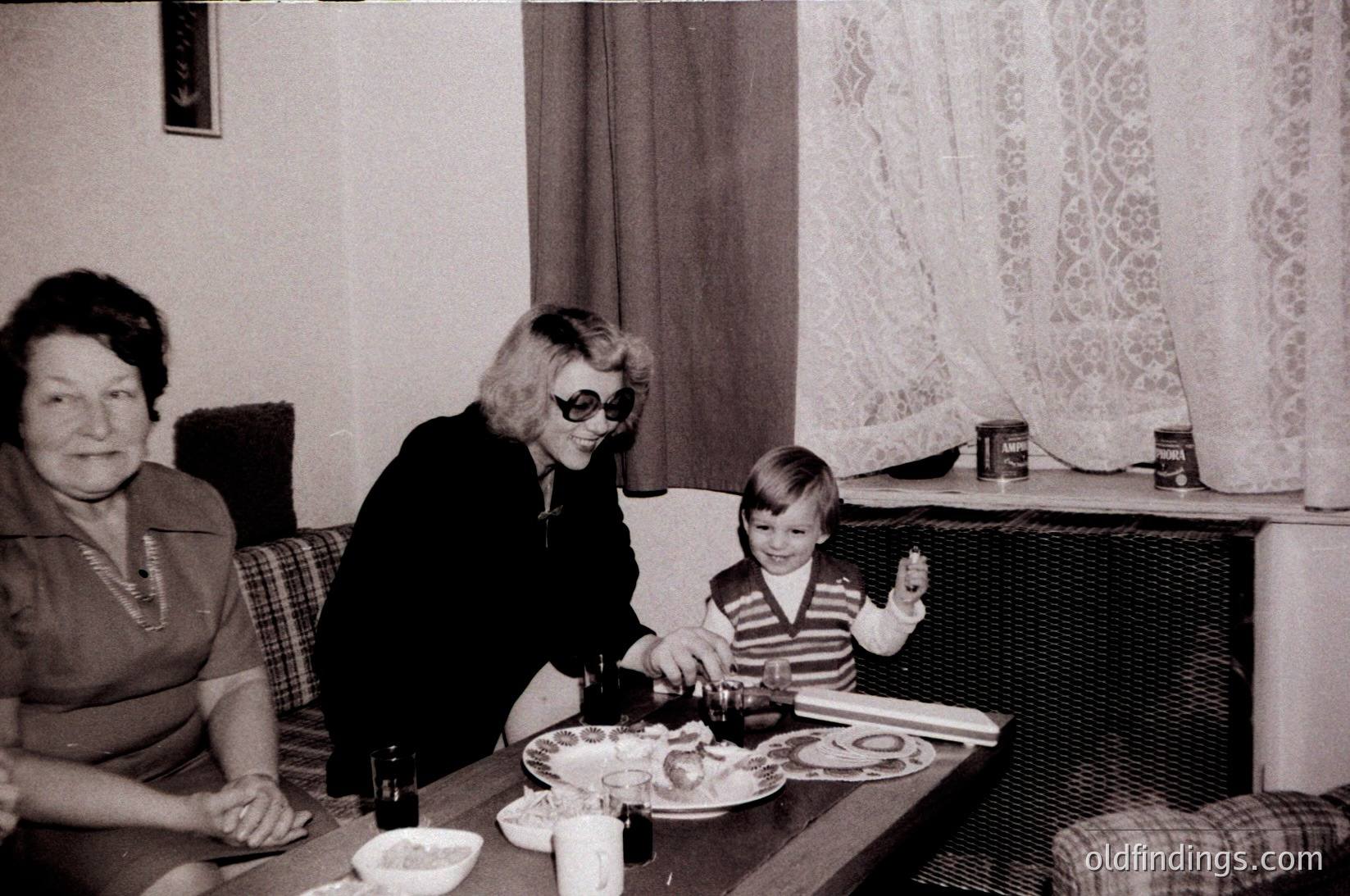 Vintage black-and-white family meal scene in a cozy 1970s living room. Three generations—elderly woman, middle-aged woman in sunglasses, and toddler—share a table with plates, glasses, and a pitcher. Checkered curtains and patterned upholstery add warmth. Candid, nostalgic moment.