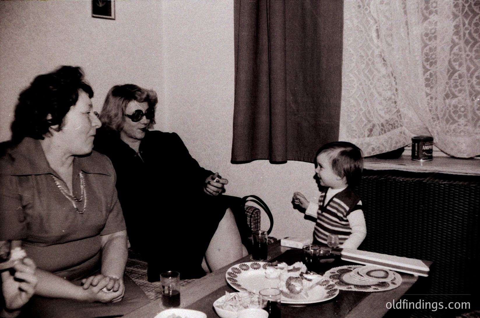 Three generations share a meal in a mid-century indoor setting—two women in formal attire (one with sunglasses indoors) and a child in a striped shirt. A vintage radio or speaker sits on the shelf, alongside a canned drink. Decorative curtains and patterned wallpaper suggest a 1960s–1970s domestic interior.