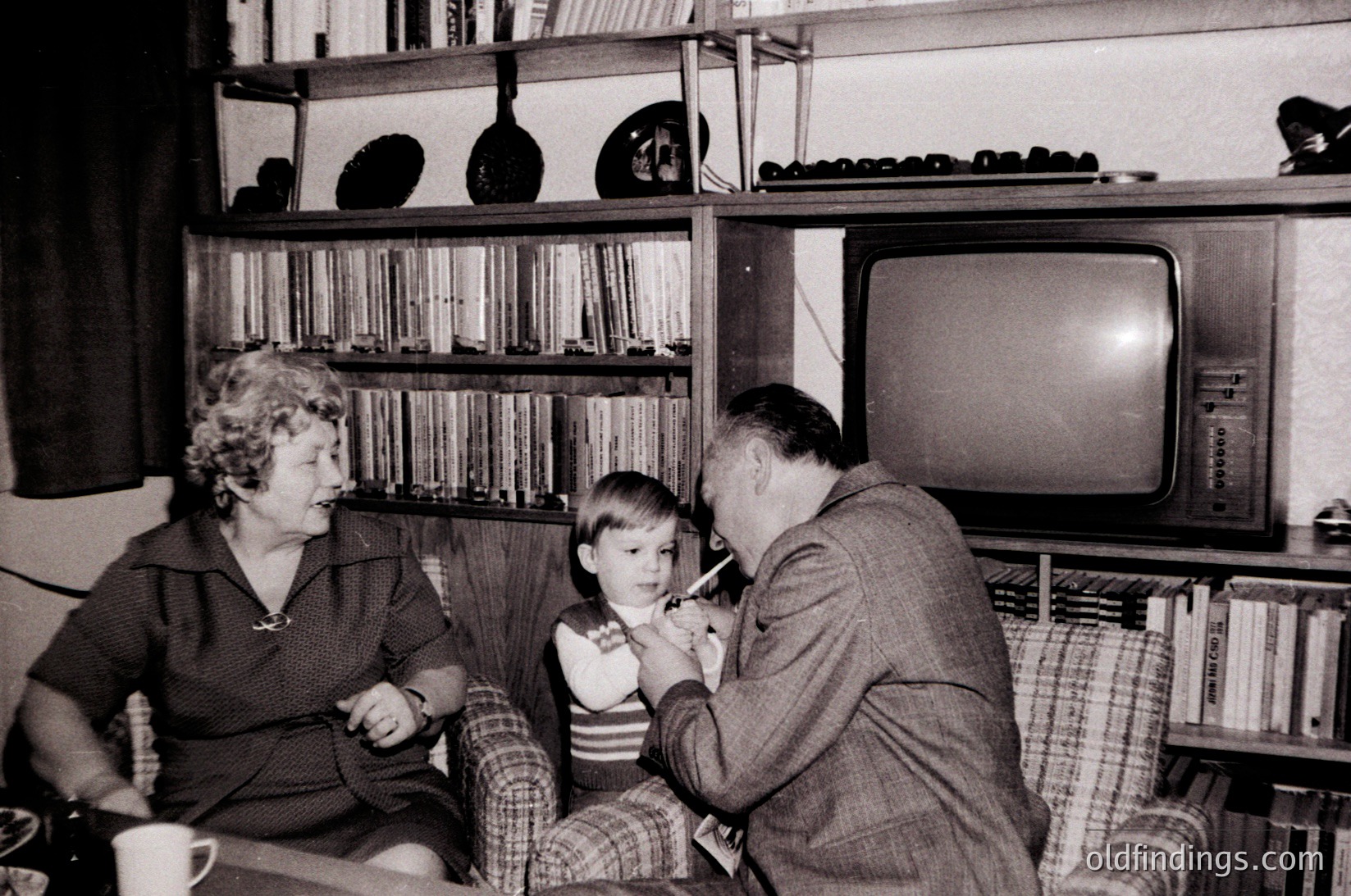 Vintage black-and-white interior shot of a mid-century living room. A man in a suit assists a young child with brushing teeth, while a woman in a dress observes. Shelves behind them hold vinyl records and books; a bulky CRT TV sits on a side table. Mid-20th century domestic life captured.