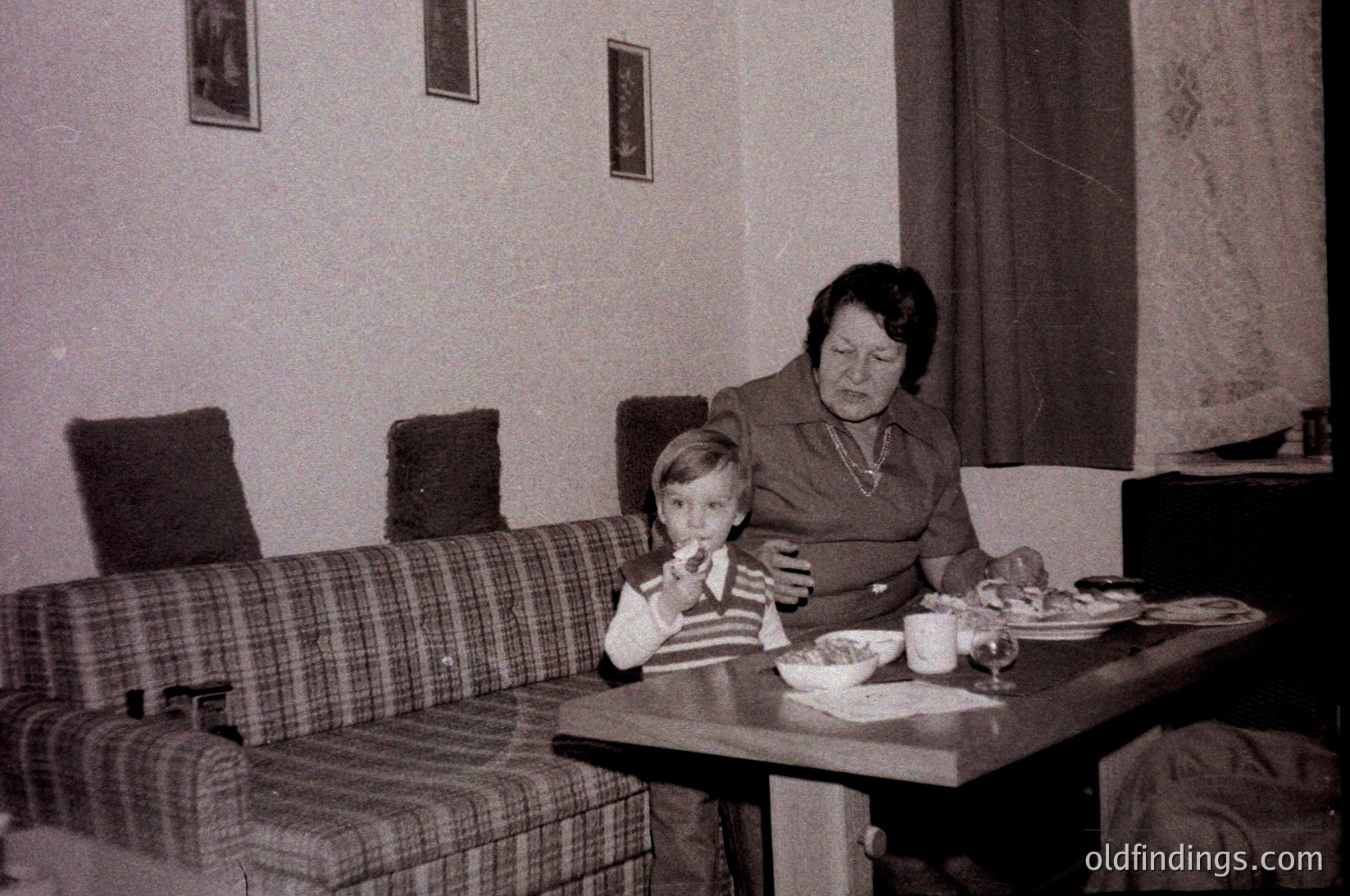 Vintage indoor scene featuring a woman and young boy seated at a small wooden table in a modest living room. Plaid sofa and floral wallpaper suggest mid-20th century decor. Table holds a bowl, plate, and glassware. Warm, candid family moment.