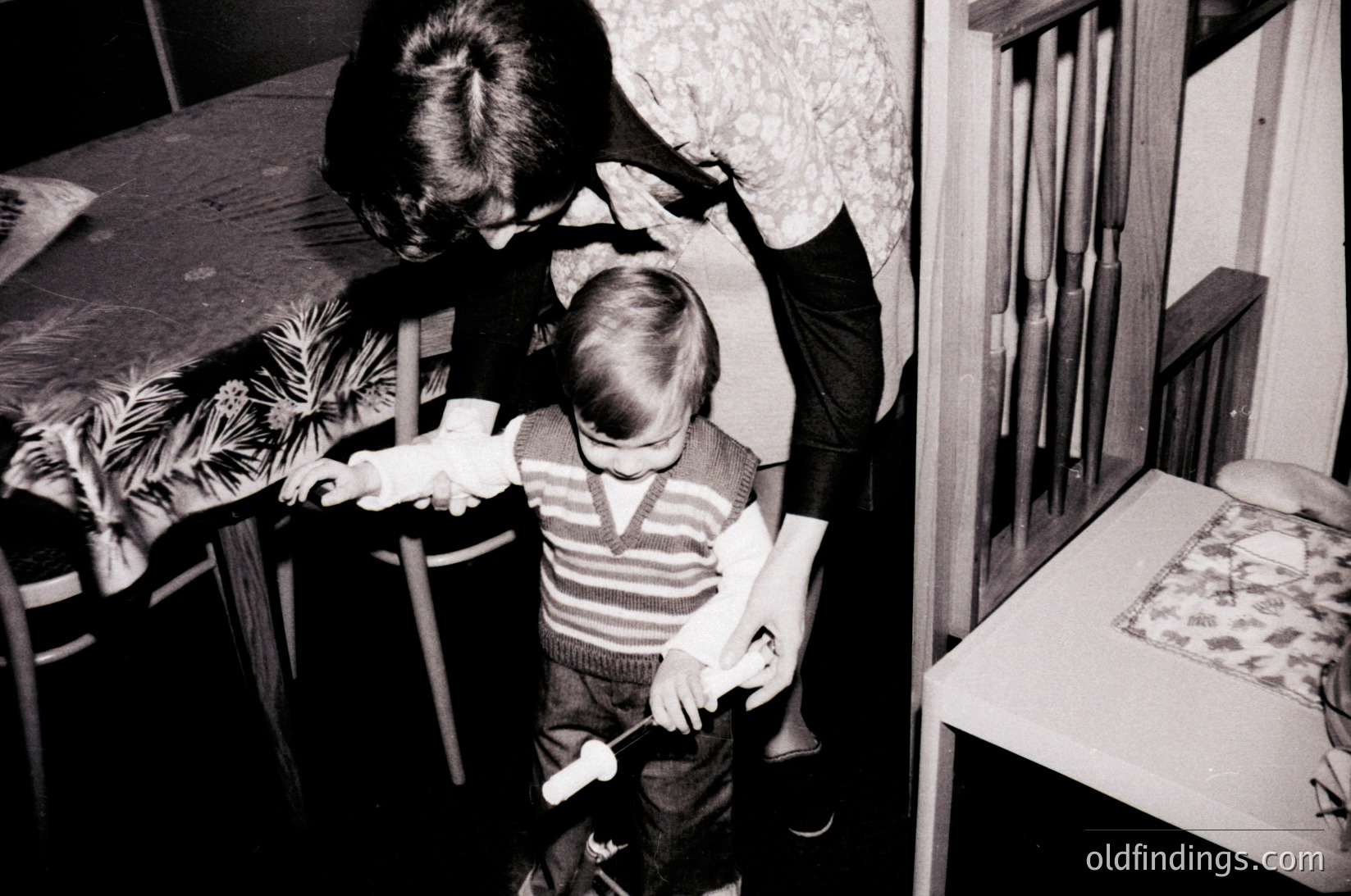 Black-and-white interior shot of a child (front) and adult (back) in a modest home, likely mid-20th century. The child stands near a wooden chair, while the adult leans over a table draped with a patterned tablecloth. Wooden furniture and a crib frame dominate the scene, suggesting a domestic setting.