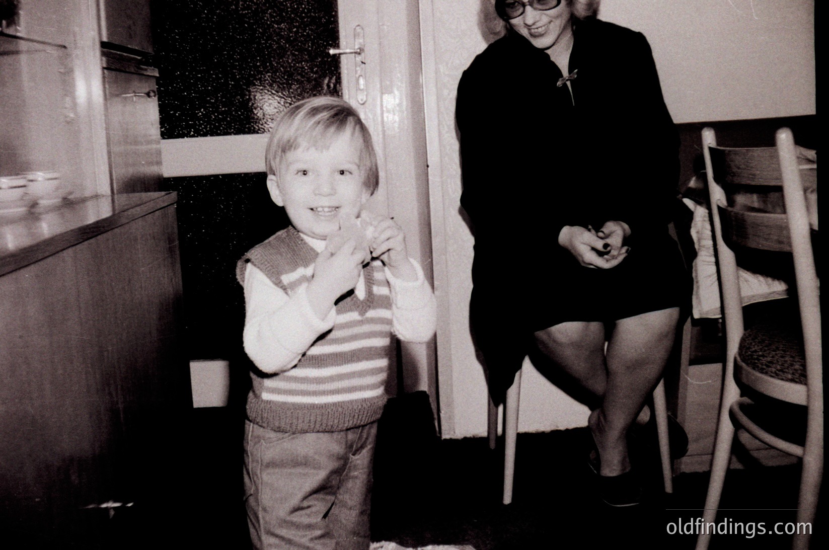 Mid-century kitchen scene: boy in striped sweater and shorts poses with toy, woman in skirt and glasses sits on stool. Wooden cabinets, vinyl window, and classic chair design suggest 1960s domestic life.