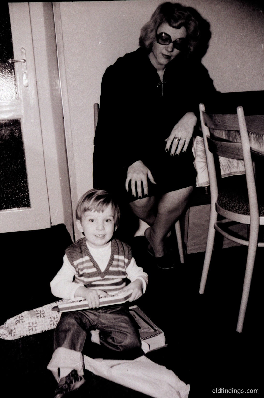 Black-and-white indoor portrait of a woman (mid-40s+) and young boy (early childhood) in a modestly furnished room, likely mid-20th century. Woman sits on a chair with a striped sweater and sunglasses indoors, boy wears a striped sweater and holds a toy. Wooden chair legs and patterned floor mat visible.