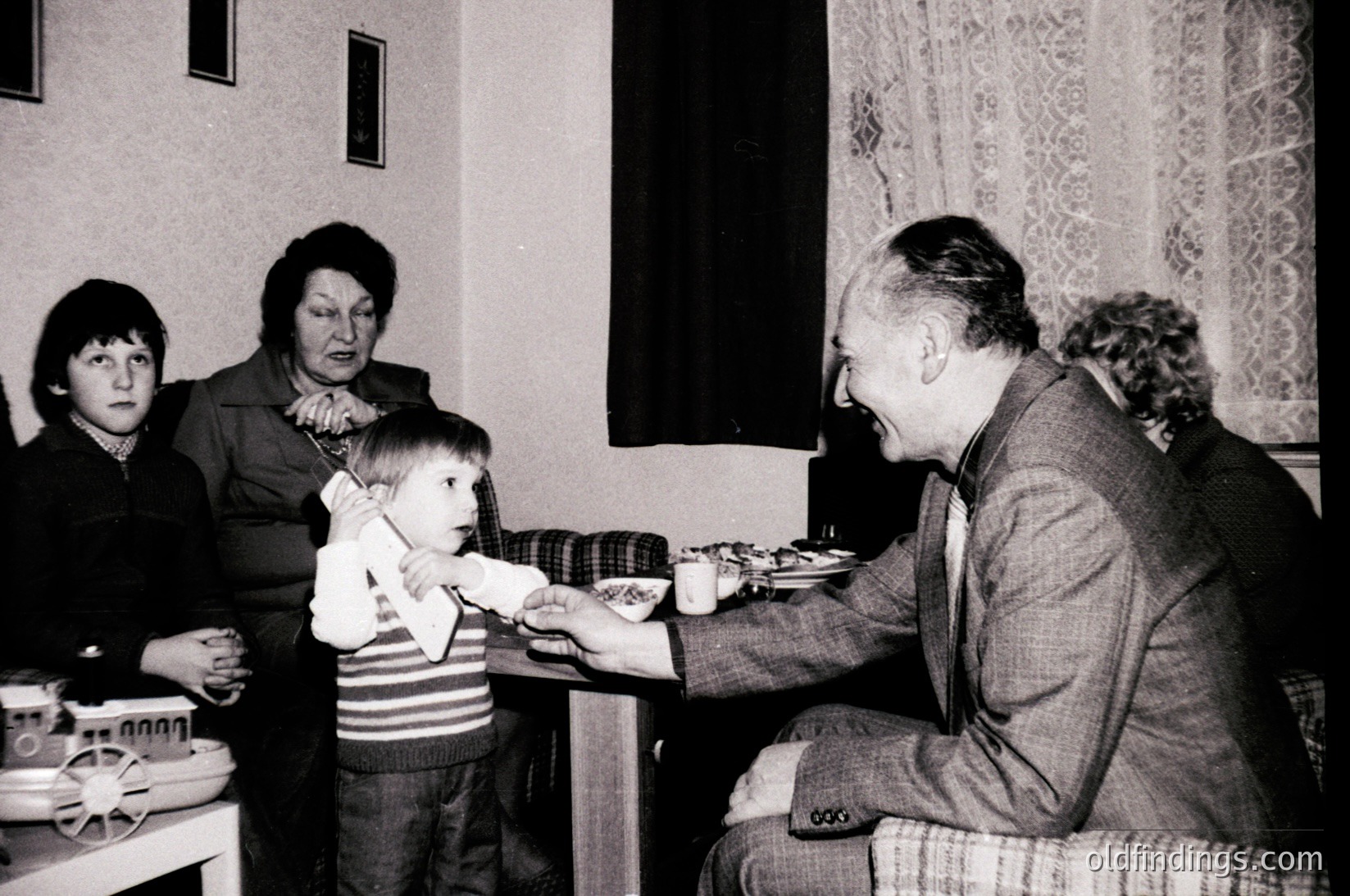Intergenerational gathering in a modest indoor setting, likely mid-20th century. A seated elderly man in a suit and striped sweater vest feeds a young child from a plate, while two women observe. Table features simple ceramic dishes and a vintage toy. Floral curtains and patterned wallpaper suggest a domestic, possibly Eastern European home.
