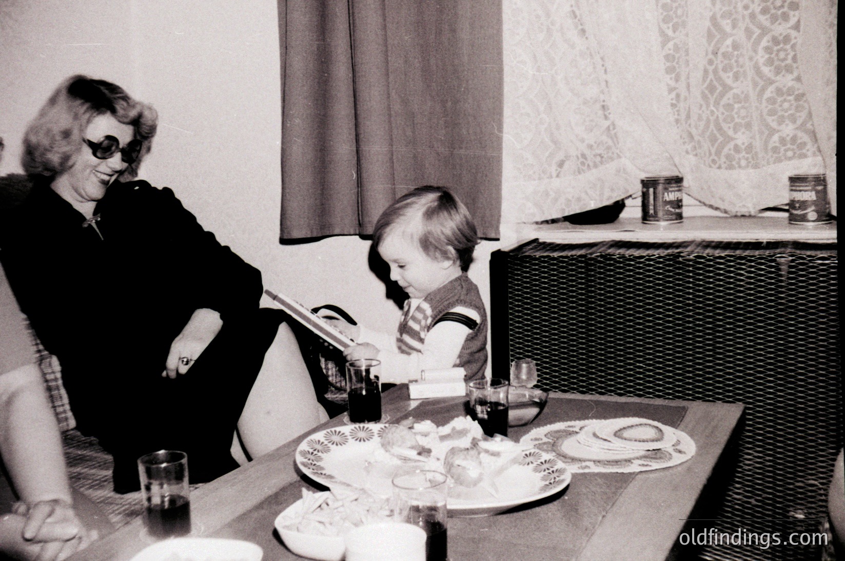 A mid-20th-century indoor scene captures a woman in sunglasses reading beside a child seated at a table. The table holds vintage plates, glasses, and a canned food item, suggesting a casual meal. Floral-patterned curtains frame the window. [Vintage 1960s-70s indoor family moment: woman reading, child seated at table with retro plates and canned goods. ]
