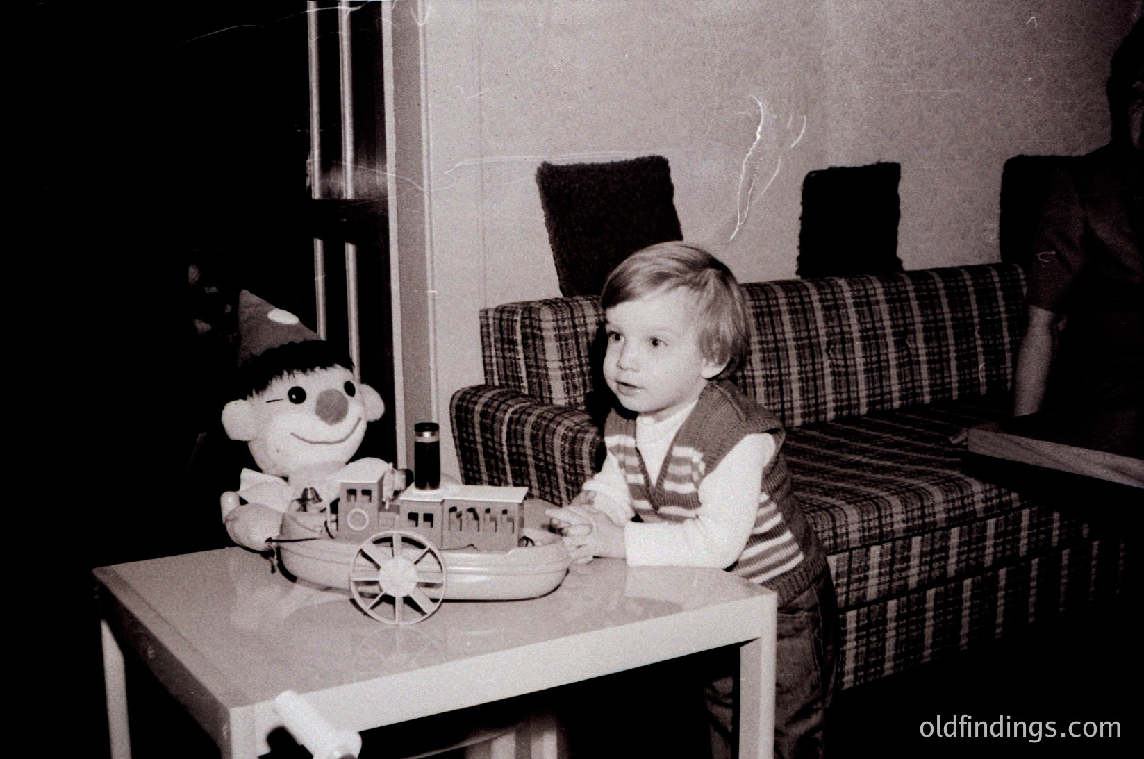 Vintage black-and-white photo of a young child seated at a small table, playing with a toy train set featuring a smiling character doll. Plaid upholstered armchair and worn carpet suggest mid-20th century domestic setting.