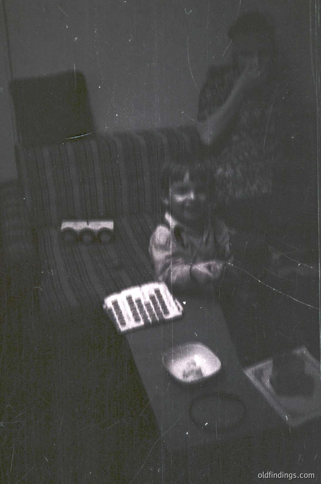 Vintage black-and-white photo of a child playing with a toy train set on a striped sofa. Reflections and scratches obscure clarity. Mid-20th century domestic interior, likely 1950s–1960s.