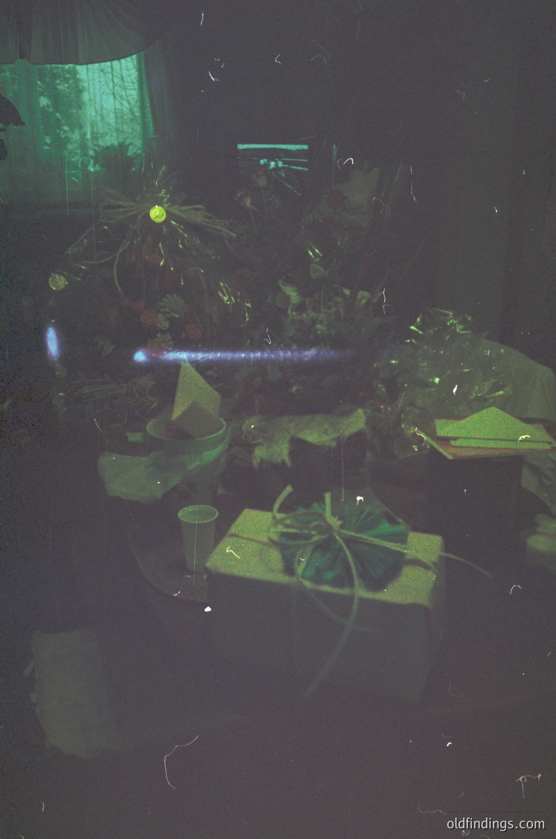 Vintage indoor scene with low-light photography, featuring a cluttered tabletop under fluorescent lighting. Green-tinted glassware, potted plants, and wrapped gifts suggest a festive or casual gathering. Reflections and lens flare add artistic distortion.