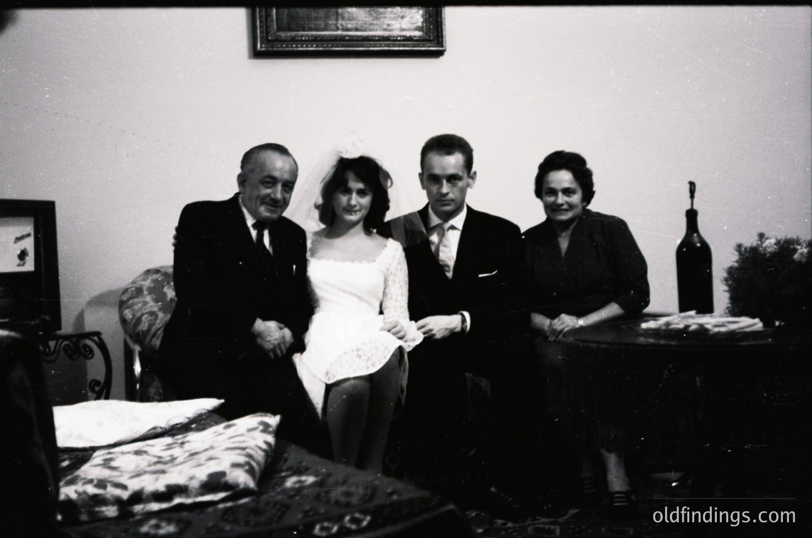 Black-and-white indoor portrait of four individuals in formal attire, likely a wedding or celebratory event. The bride wears a fitted dress with a veil, flanked by a man in a suit and tie and a woman in a dark blazer. The groom, also in a suit, stands beside her. A framed picture hangs above, and a wine bottle rests on a side table with floral-patterned upholstery.