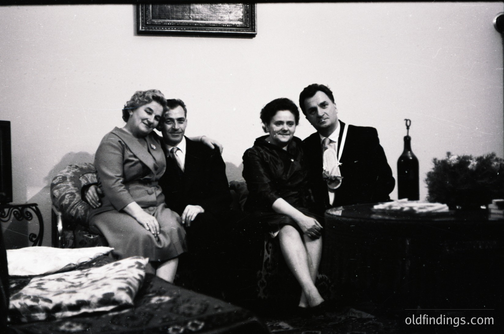 Four individuals pose indoors in formal attire, likely 1950s–1960s. Men wear suits with ties; women in structured dresses. Bottle and glass suggest a social gathering. Plain walls, patterned sofa, and framed artwork hint at mid-century domestic decor.