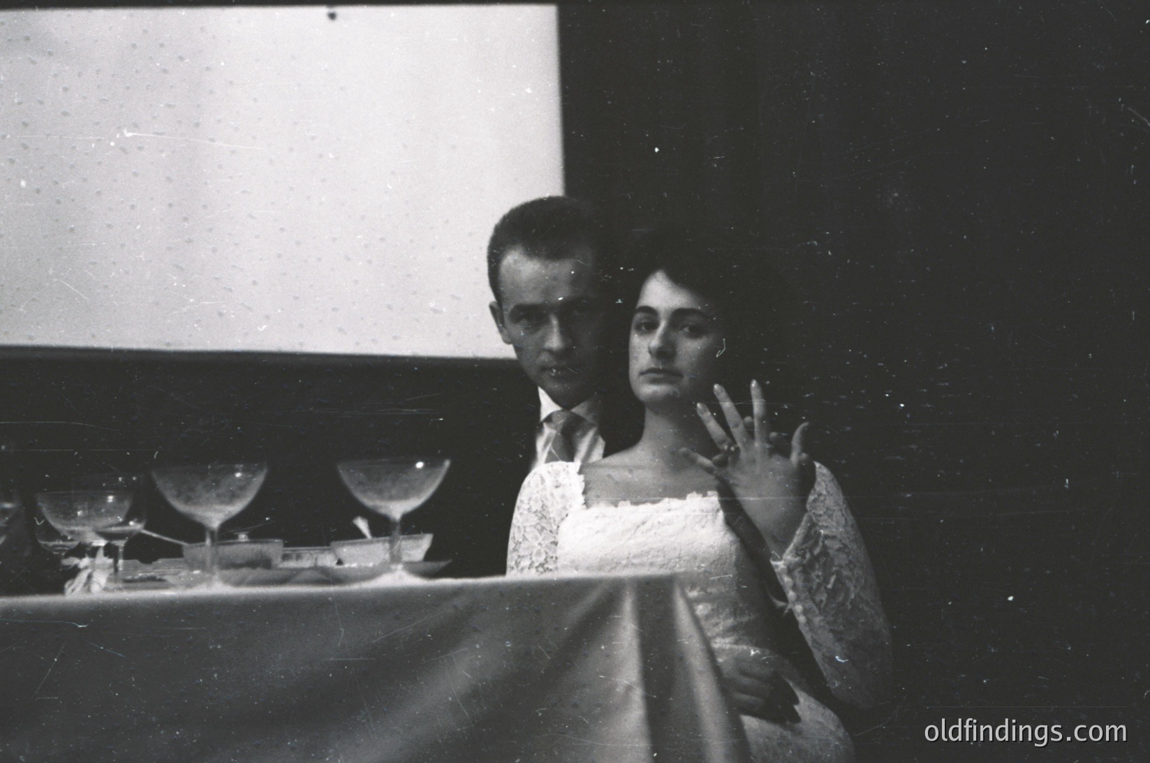 Couple seated at a table in a dimly lit, speckled projection room, likely a 1950s–1960s cinema. Man wears a dark suit; woman in a lace-trimmed dress, glasses, and a hand gesture. Empty wine glasses and a plate with remnants suggest an intermission. Vintage film grain and lighting evoke mid-century social culture.
