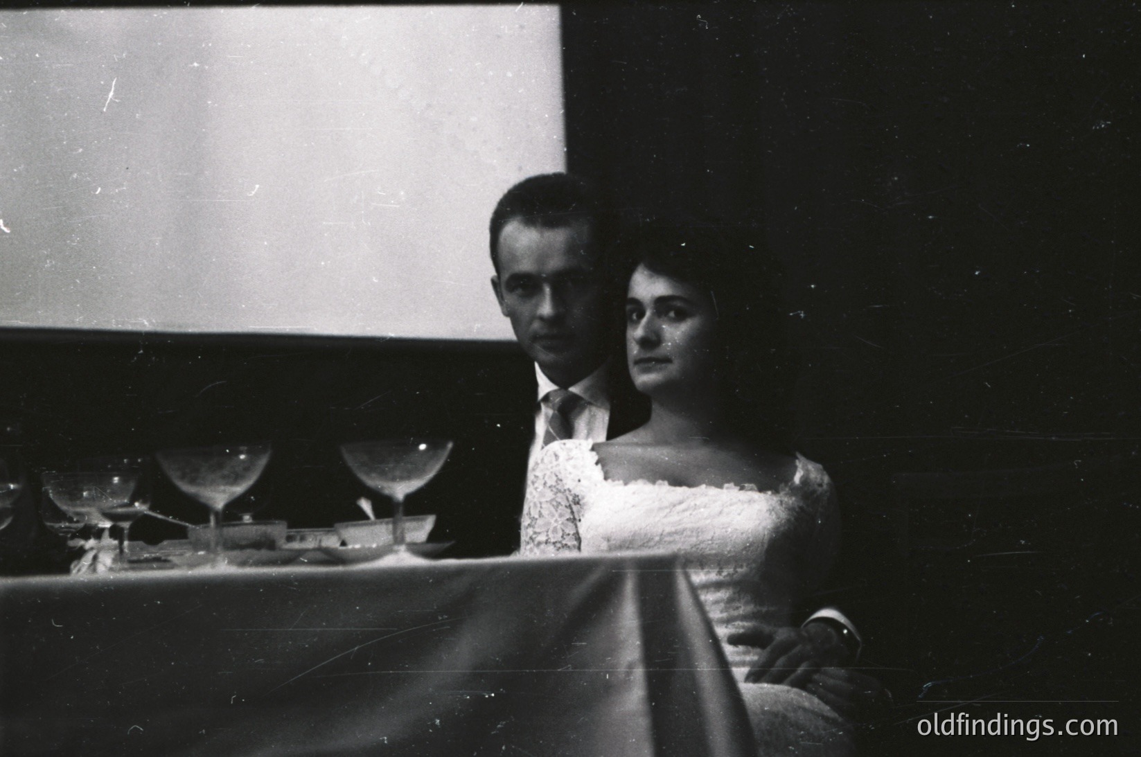 Black-and-white portrait of a man and woman seated at a piano, likely from the 1940s–1960s. The man wears a suit with a white shirt, while the woman dons a formal dress with a fur collar. Reflections in the piano’s polished surface reveal a mirror and chandelier, suggesting an elegant indoor setting. Ideal for vintage music, formal events, or historical research.