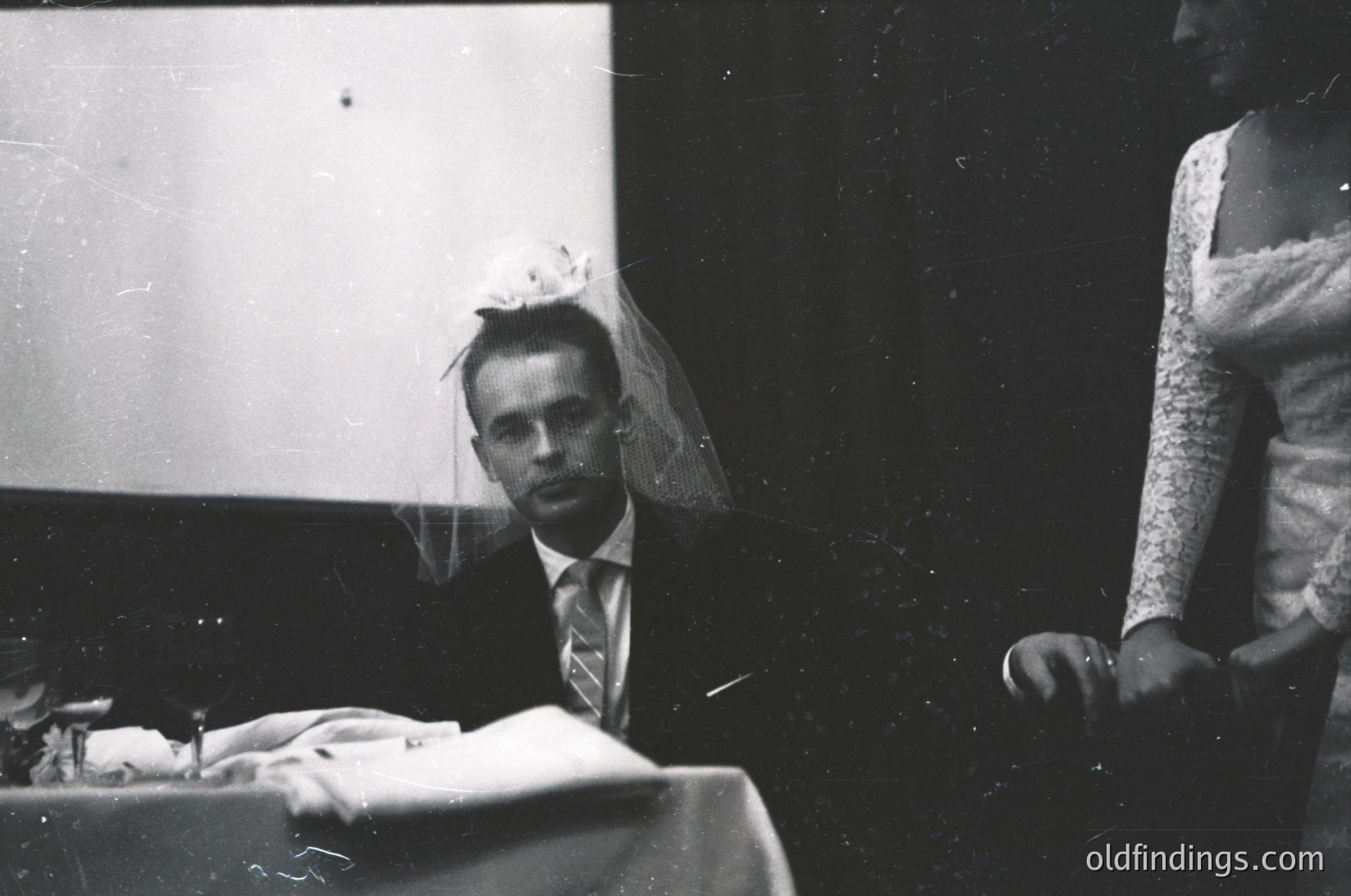 Black-and-white portrait of a groom in a tuxedo with veil, seated at a table with a white cloth. Partial view of bride in lace dress beside him. Formal indoor setting, likely a 1950s-1960s wedding. Groom’s expression is neutral, framed by vintage lighting.