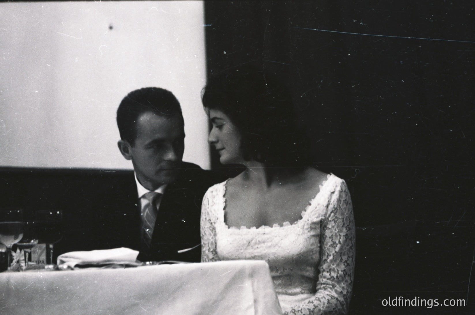 Couple seated at a formal table, likely at a 1960s-1970s wedding or gala. Man in dark suit, woman in lace wedding dress. Indoor setting with dim lighting and draped tablecloth.