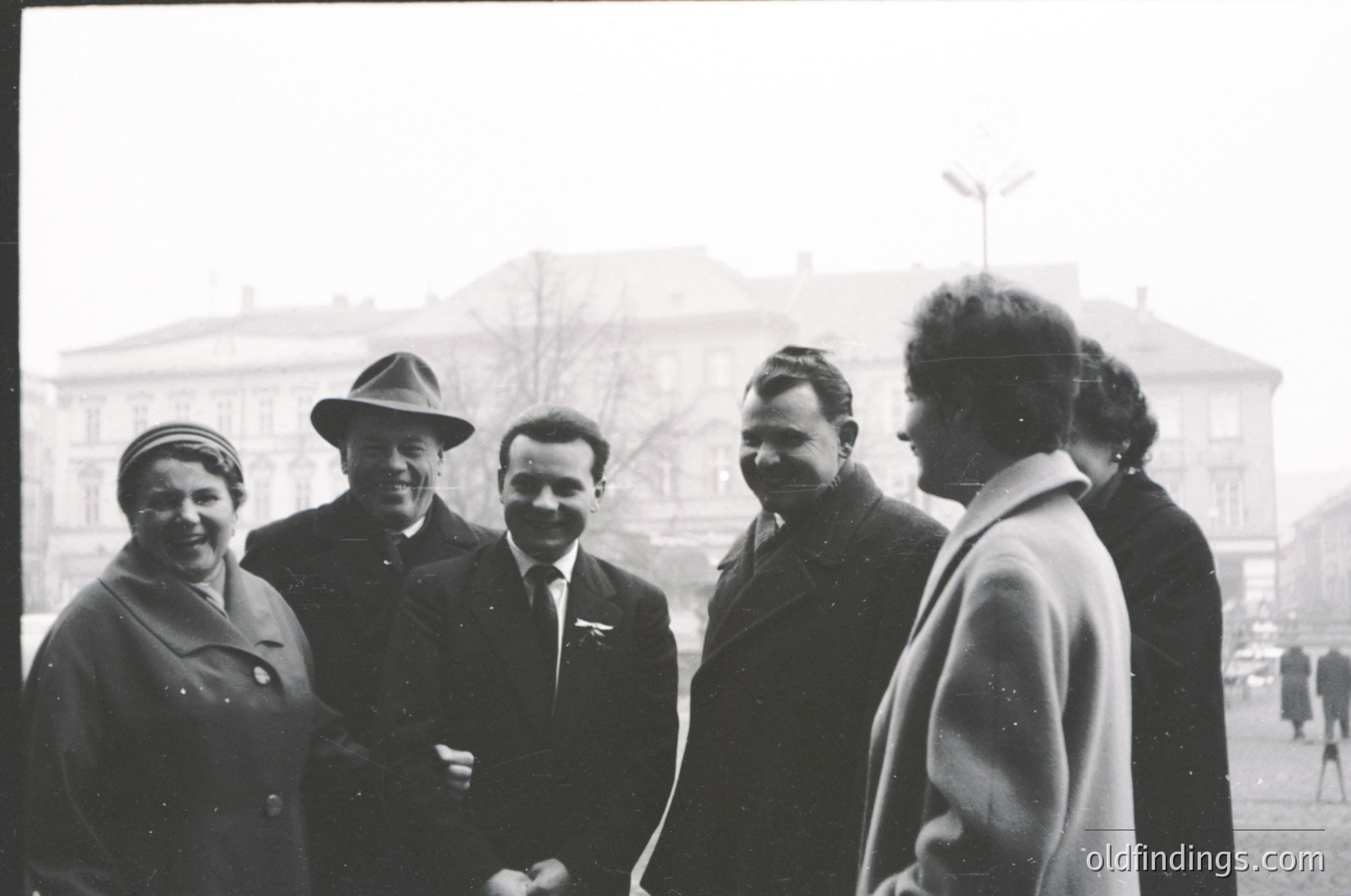 Five individuals pose outdoors in 1960s Eastern Bloc attire—men in suits with badges, women in coats and hats—against a Soviet-style building. Classic black-and-white street photography captures mid-century camaraderie.