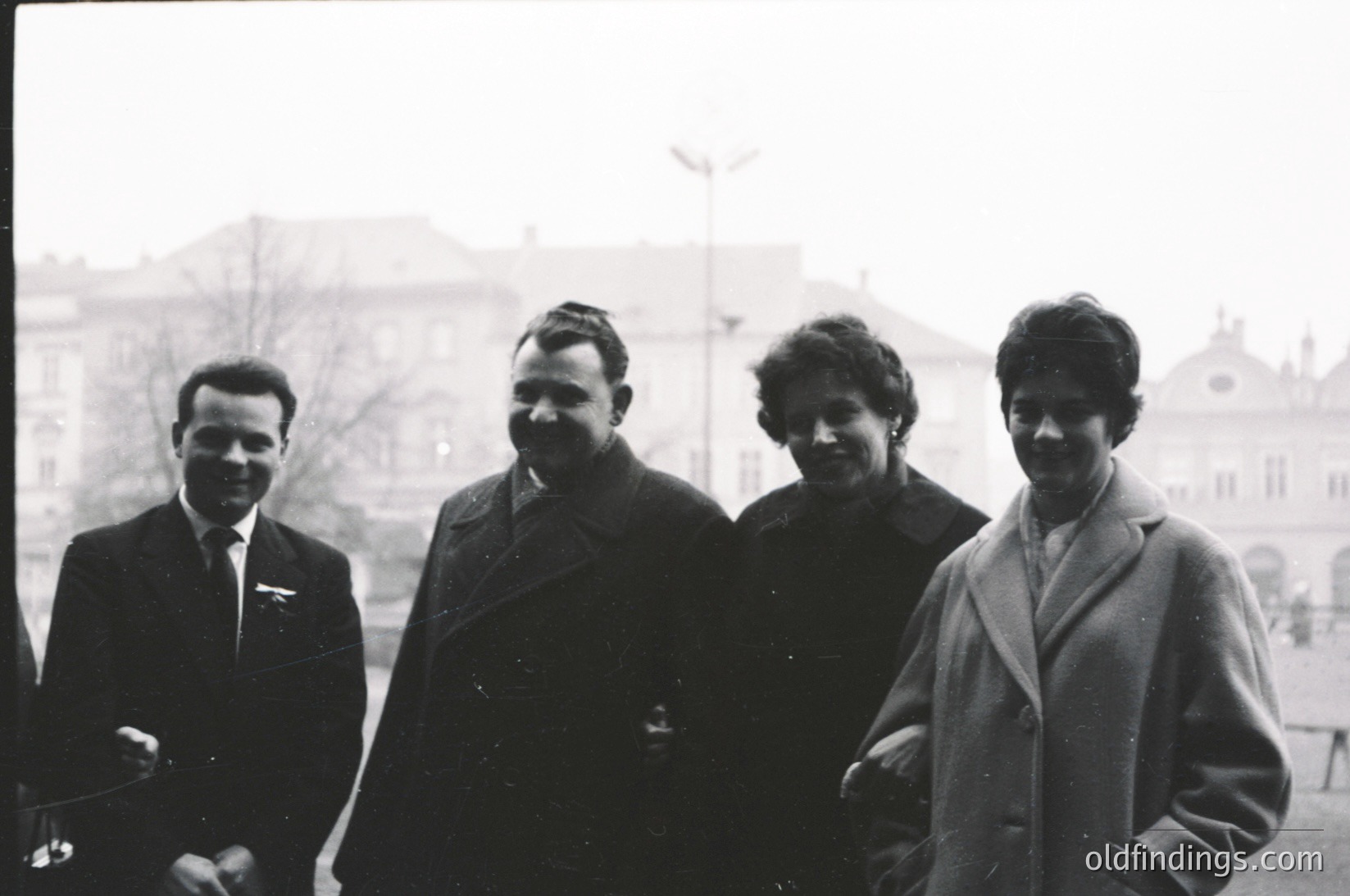 Four individuals pose outdoors in mid-20th century attire, likely 1950s–1960s. The man on the left wears a pilot’s jacket with an insignia, suggesting aviation or state-sponsored travel. The central figure’s overcoat and hairstyle reflect Eastern Bloc fashion. The background features Soviet-style architecture with blocky, utilitarian design. Urban setting, possibly Eastern Europe.