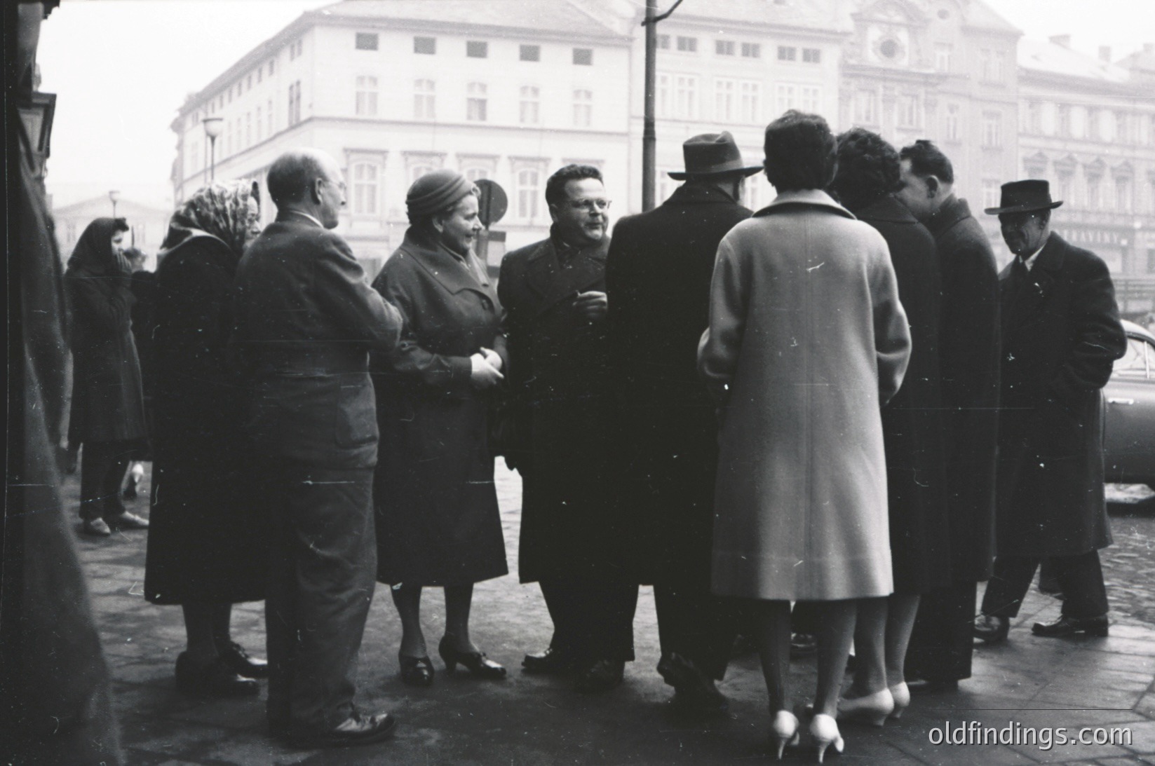 Group of 12+ men and women in formal 1940s attire—overcoats, hats, gloves—standing outdoors near a grand building with classical architecture. Central figures appear to be engaged in conversation. Likely European urban setting, possibly post-WWII era.