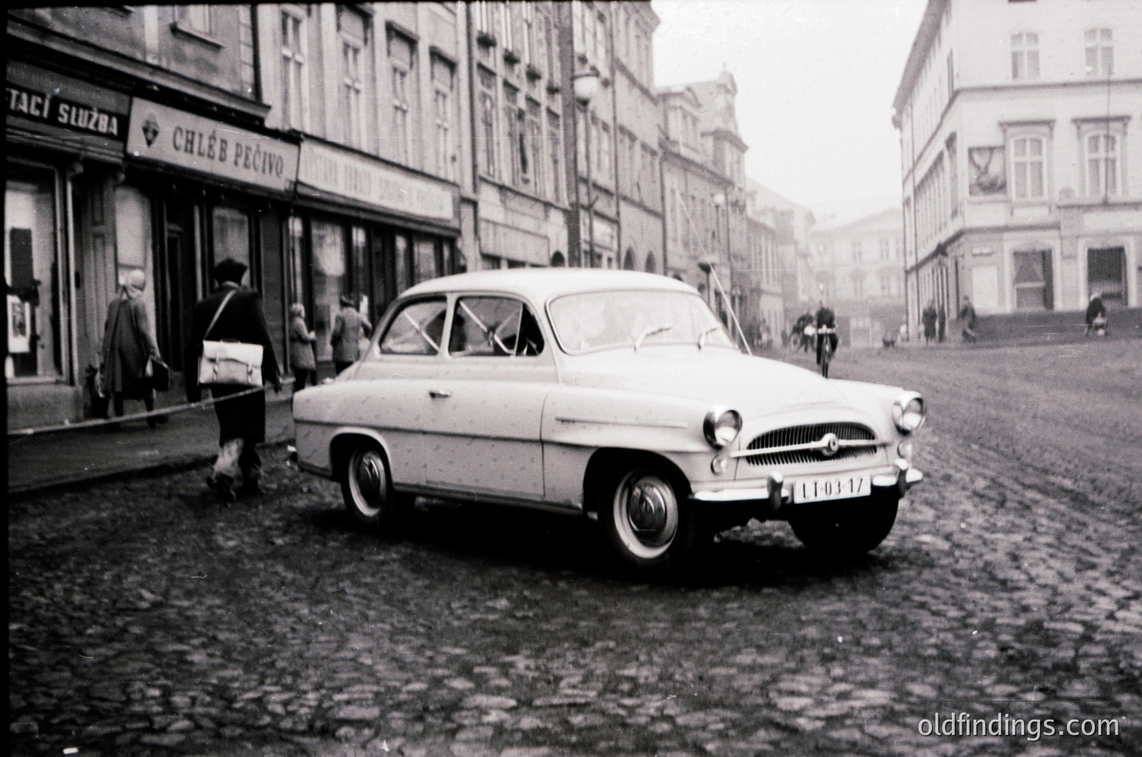 Vintage 1950s-era Škoda car parked on cobblestone street in European city. Signs in Czech ("Chléb Pečivo") indicate bakery. Pedestrians in mid-20th-century attire. Classic urban architecture with multi-story buildings.