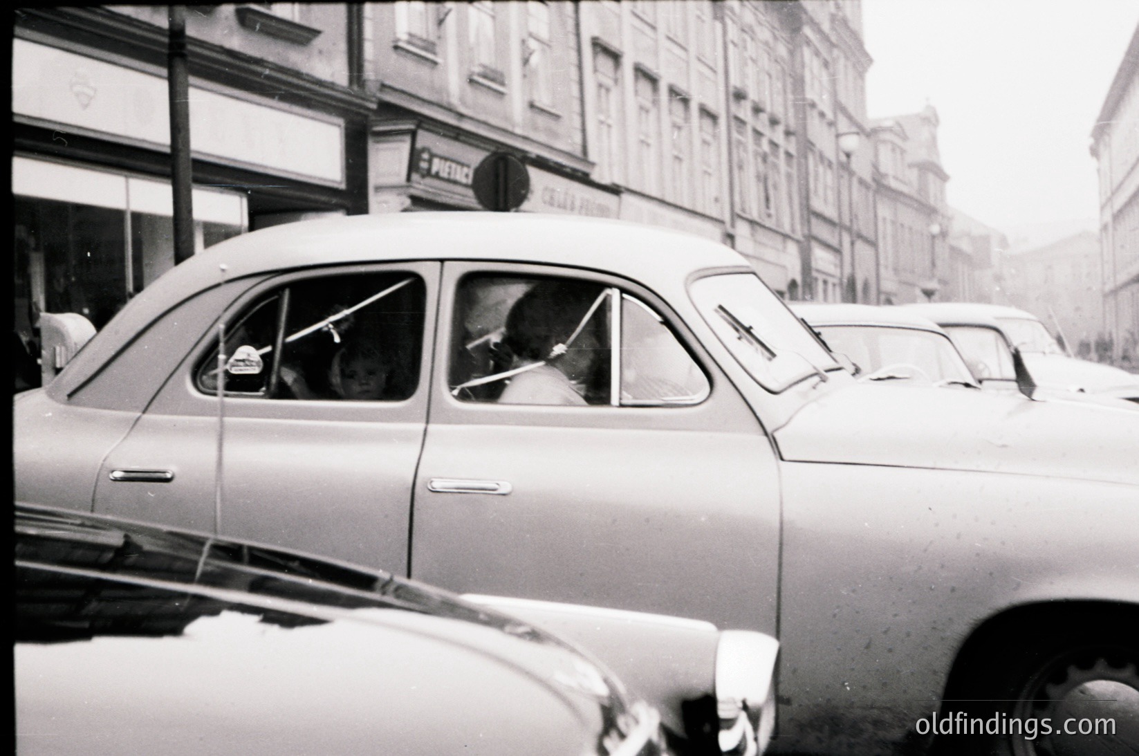 Classic mid-century taxi with "POLICE" sign on roof, likely a vintage Škoda or similar Eastern Bloc model. Urban street scene with multi-story buildings and commercial signage. Black-and-white, suggesting 1950s–1970s timeframe. Distinctive retro urban aesthetic.