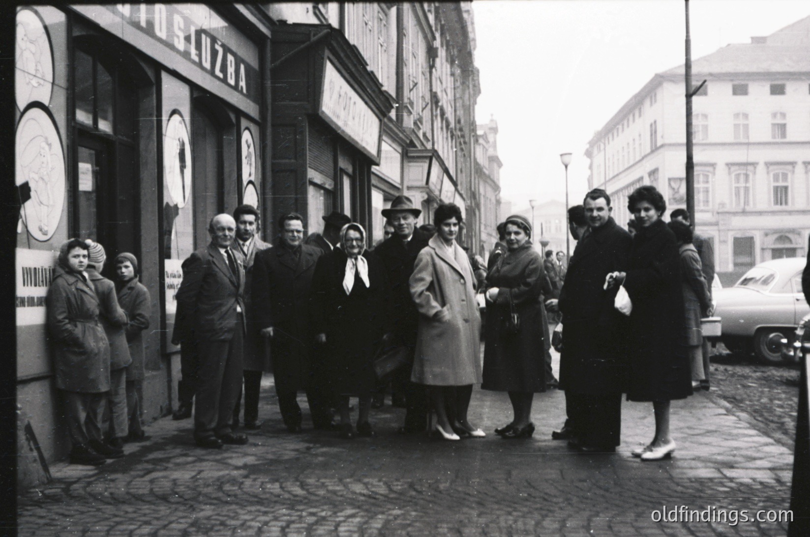 Vintage group portrait on a European city street, likely 1950s–1960s. Nine individuals in mid-century attire—coats, hats, and structured suits—pose outside a storefront with a clock and signage in Cyrillic. Cobblestone pavement and parked vintage cars suggest a small-town or urban setting.