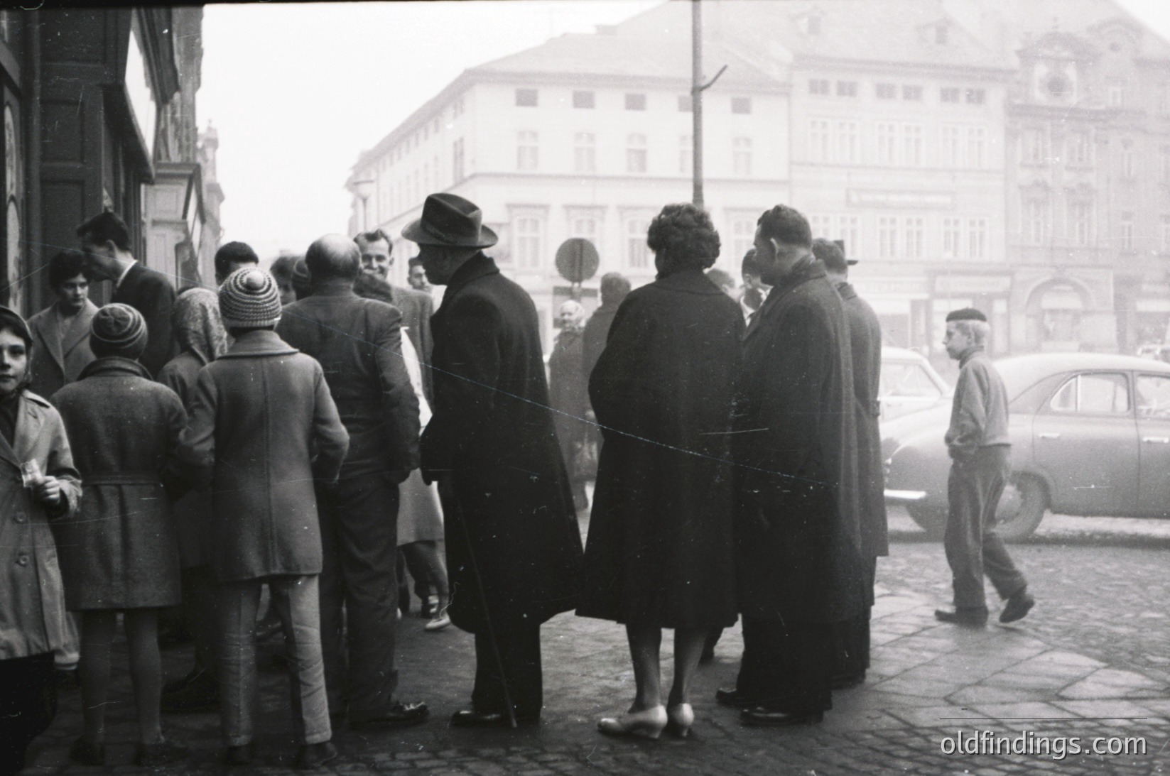 A mid-20th century street scene featuring a diverse crowd in winter attire, lined up outside a building with ornate facades. Men in suits, hats, and women in coats and hats suggest a formal or official gathering. A vintage car parked nearby hints at mid-century urban life. Foggy atmosphere enhances the historical ambiance.