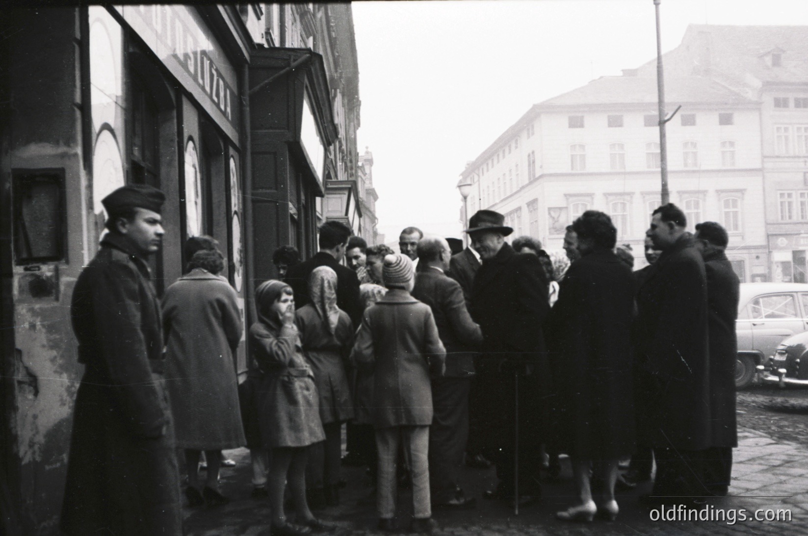 A mid-20th century street scene with a diverse crowd gathered outside a building with peeling paint. Men in suits, women in coats, and children in school uniforms stand in a queue, likely waiting for services or supplies. Classic cars and Soviet-era architecture hint at Eastern Europe, possibly or . Urban life and rationing-era daily life captured.