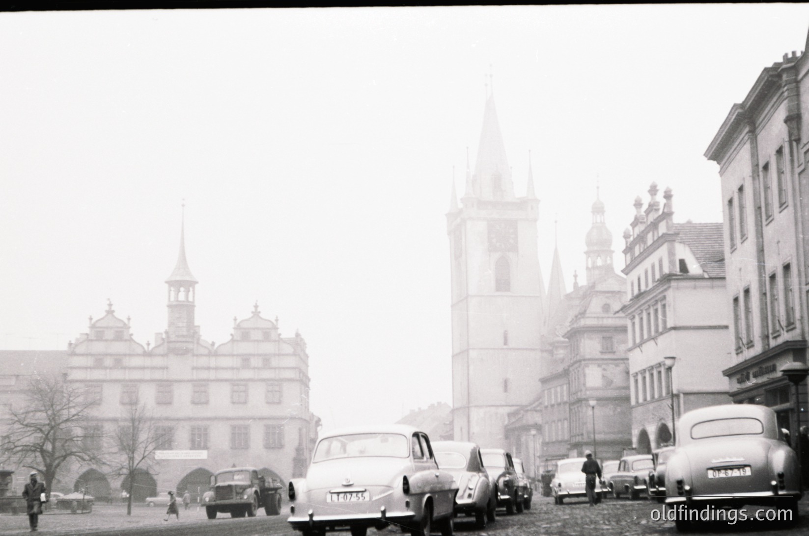 Mid-20th century European cityscape with misty fog obscuring details. Classic Baroque-style buildings with gabled roofs and ornate facades dominate the scene. Vintage cars, including a white sedan in foreground, suggest 1950s–1960s era. Pedestrians and parked vehicles populate the cobblestone street. Prominent church tower with spire in background.