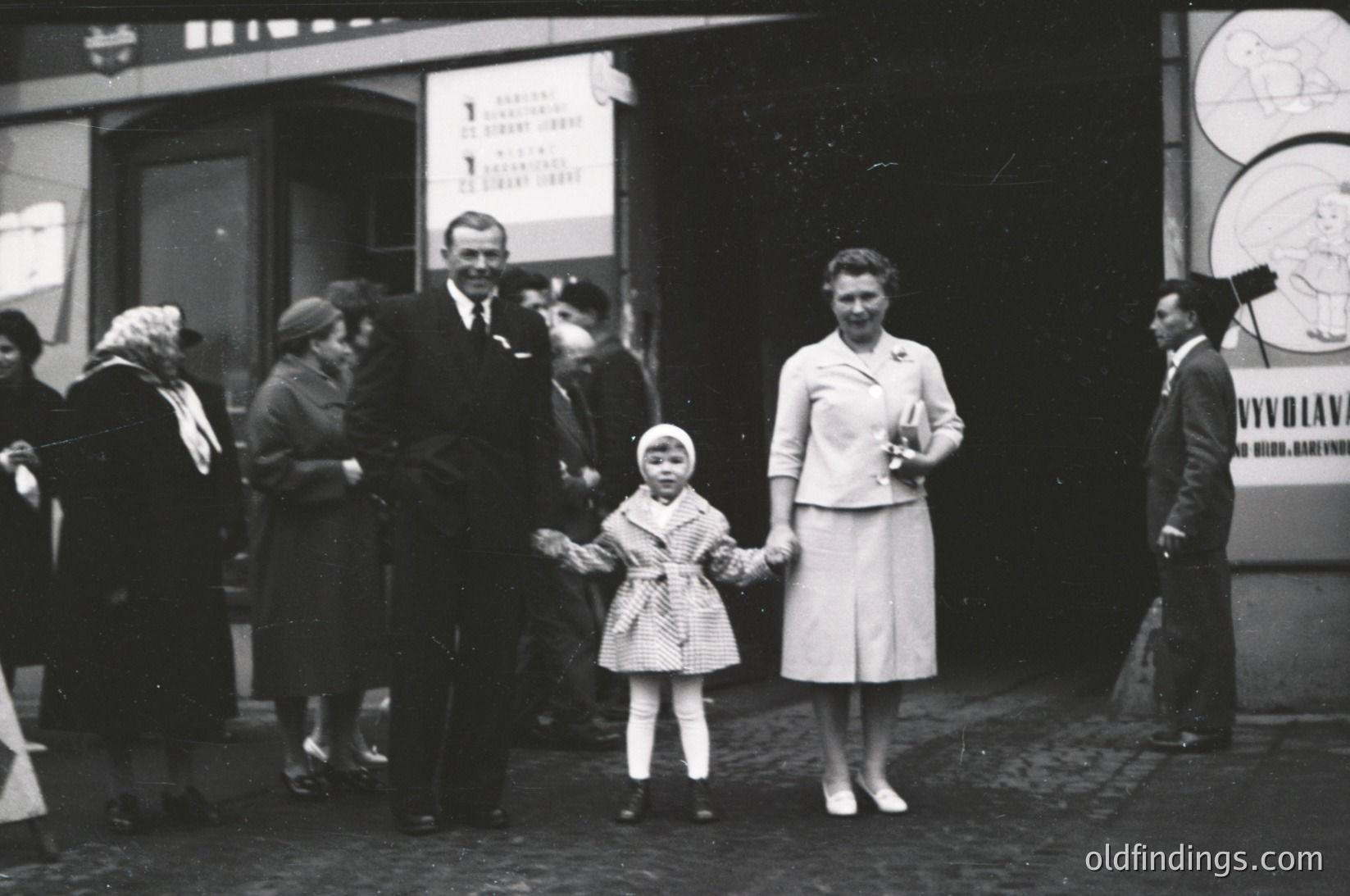 A mid-20th century street scene featuring a family posing outside a building with a sign reading "VYVOLOVÁN" (likely Czech/Slovak for "draft registration"). A woman in a structured suit holds a young girl in a polka-dot dress, while a man in a suit stands beside them. Crowd in vintage attire, cobblestone pavement, and a poster with a map in the background.