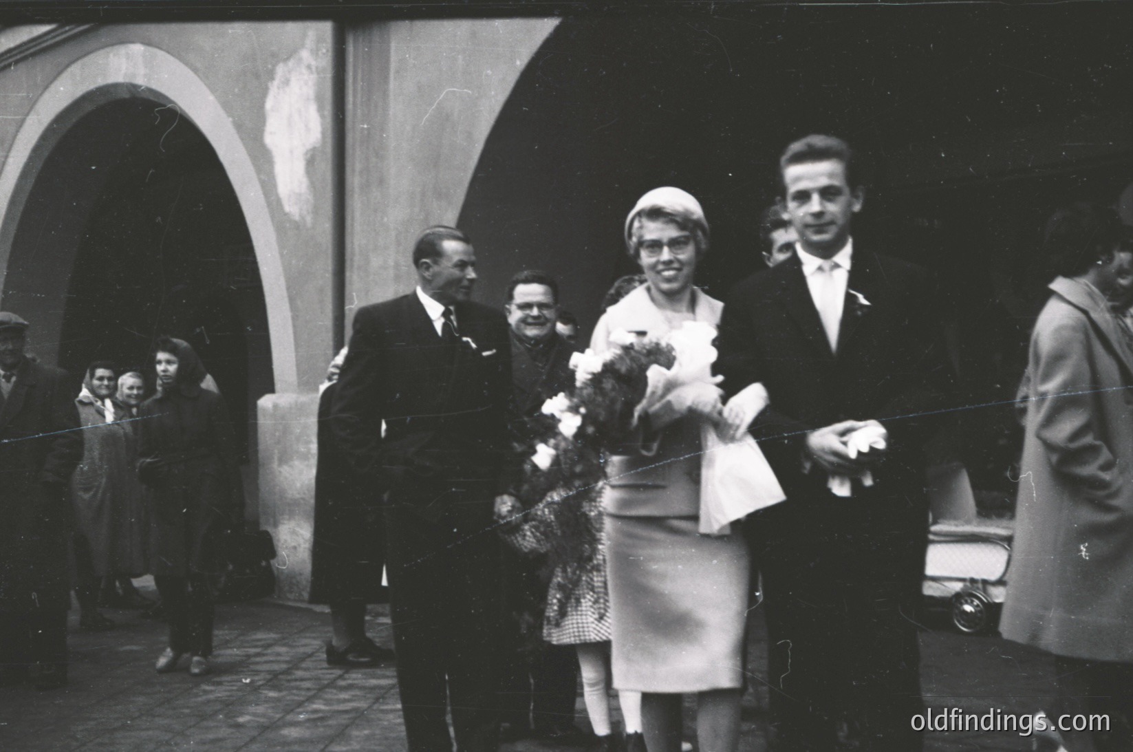 Black-and-white funeral procession outside a building with arched entryways. Central figures: woman in formal dress holding floral wreath, flanked by men in suits. Crowd in background, some holding flowers. Vintage car parked nearby. Likely Eastern European, 1950s–1960s.