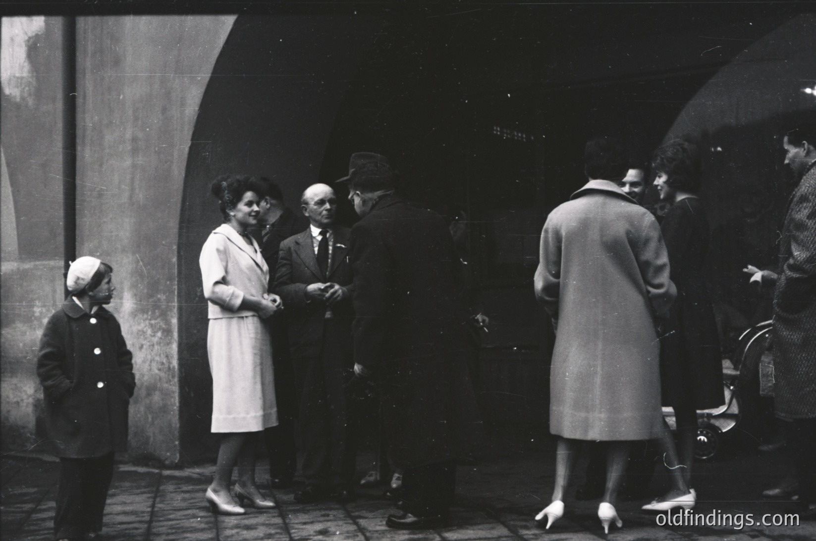 Mid-20th century street scene under arched stone passageway. Group of adults in formal attire—men in suits, women in knee-length dresses and coats—engaged in conversation. Child in white blouse and dark skirt stands nearby. Architectural details include curved stone arches and weathered walls. Likely European urban setting, 1940s–1950s.