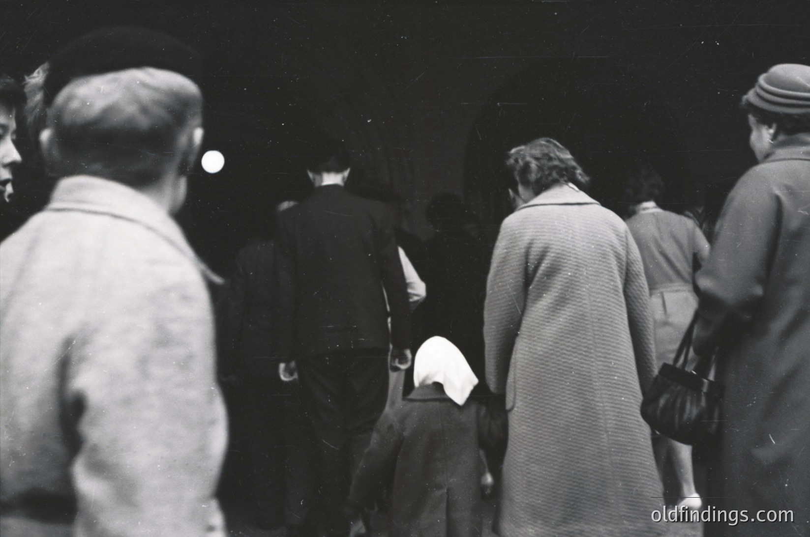 A candid black-and-white street scene from the 1950s–60s, featuring a diverse group of people in mid-20th-century attire. A woman in a long coat and headscarf leads a child, while others in hats and coats walk in a dimly lit urban area. Architectural details like a building facade and streetlights suggest a European city setting.