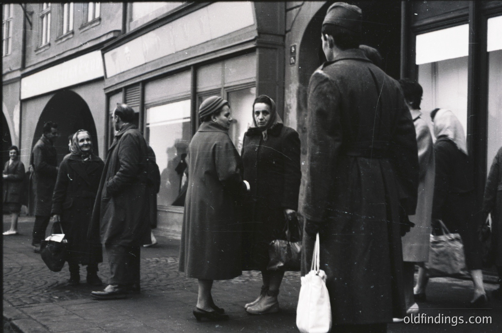 Black-and-white street scene featuring mid-20th century European urban life. Crowd of individuals in heavy winter coats, carrying bags, gathered outside a storefront with arched doorway. Architectural details include cobblestone pavement and brick buildings. Likely