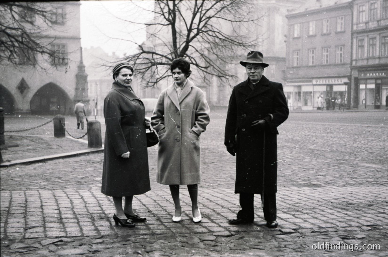 Three individuals pose on cobblestone street in 1950s European city square. Women wear knee-length coats with structured shoulders; man in overcoat and fedora. Gothic-style buildings and bare trees frame scene.